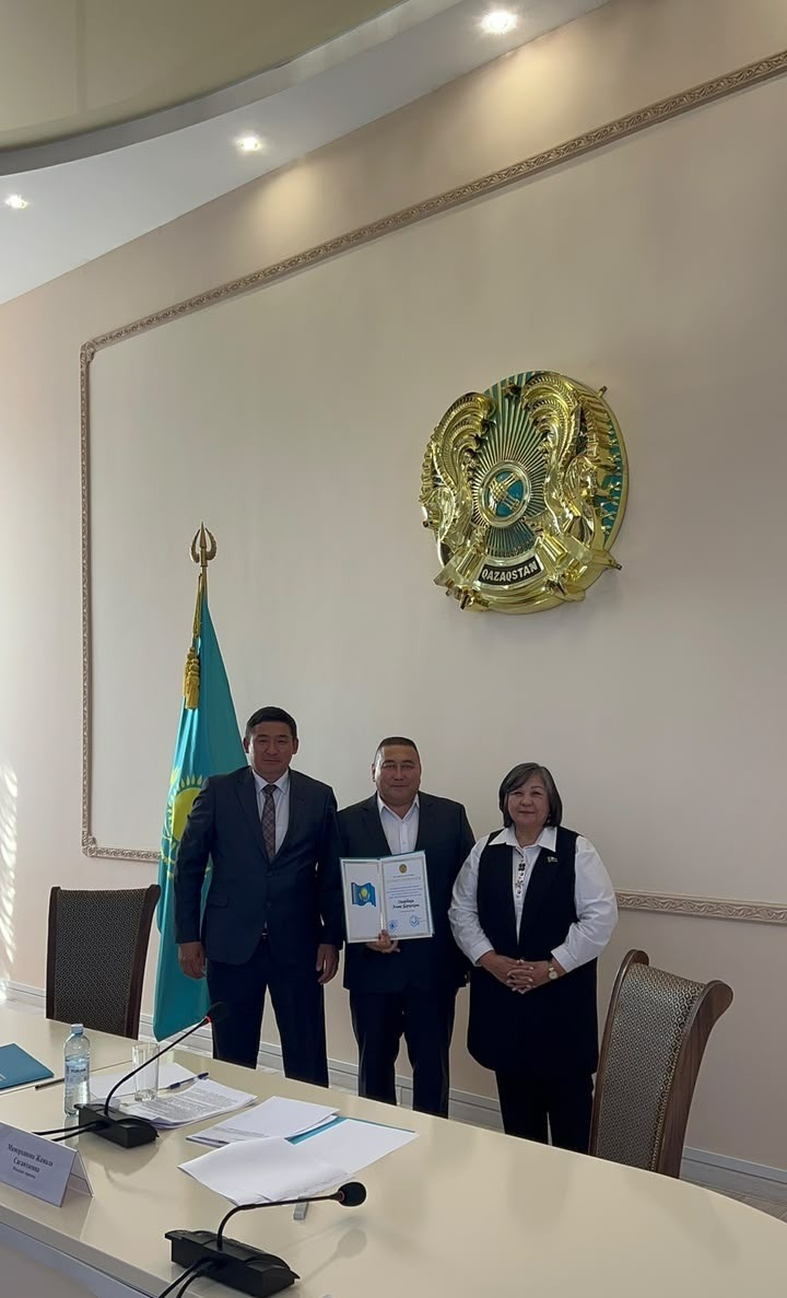 Қазақстан Республикасының мемлекеттік рәмігінің алдында тұрған үш адам. Ортадағы ер адам сертификат ұстап тұр.