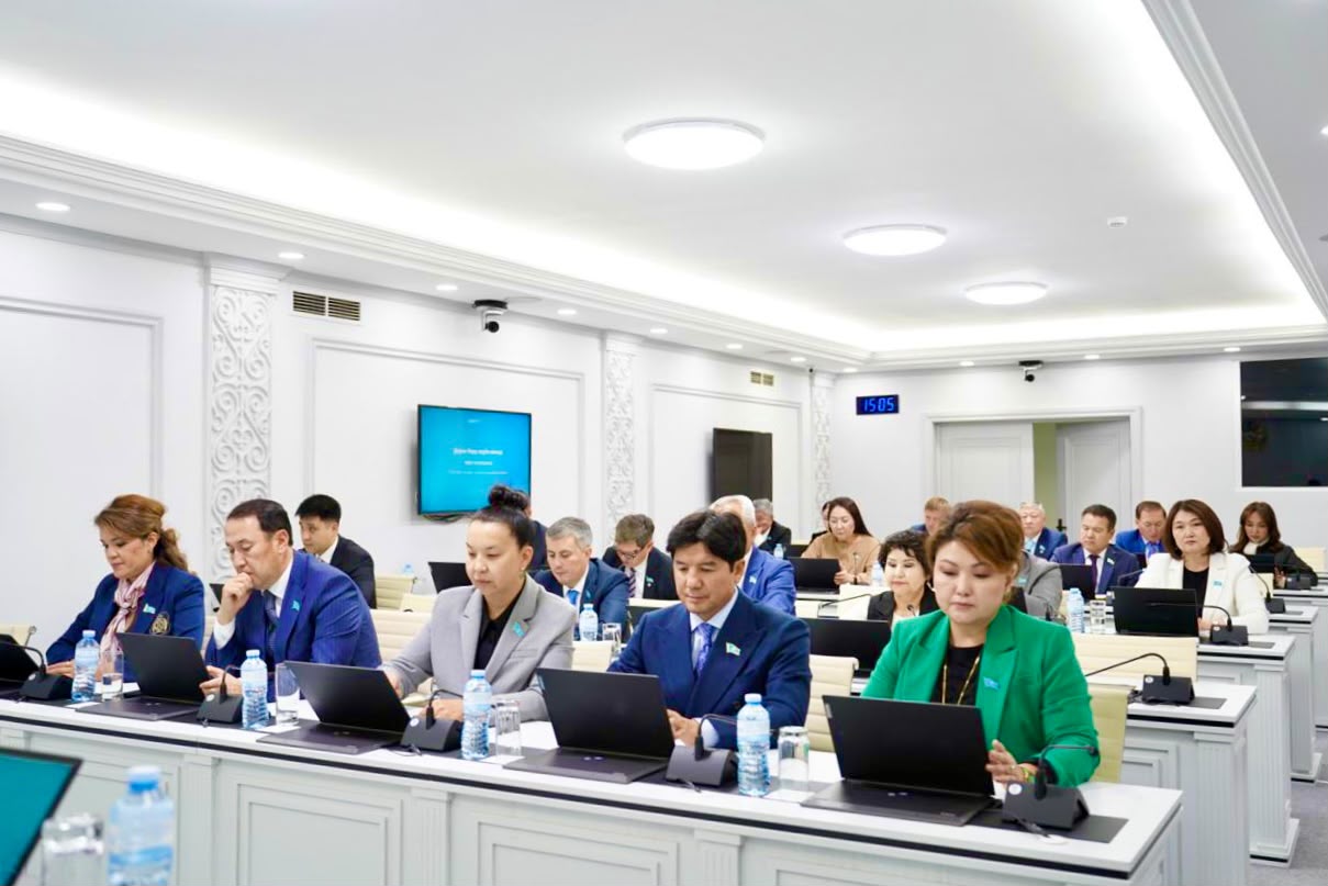 Жиналыс залында ноутбуктермен жұмыс істеп отырған ресми киінген адамдар тобы.