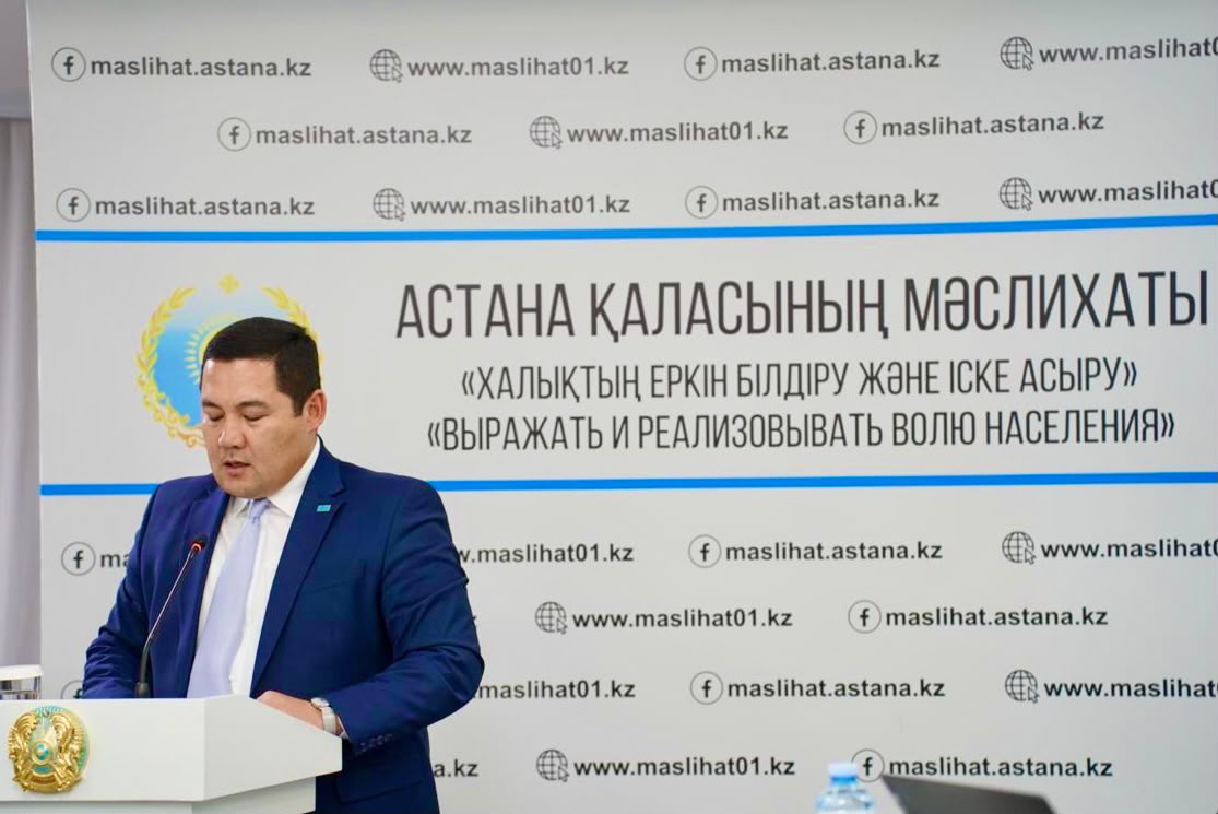 Астана қаласының мәслихатының алдында сөйлеп тұрған көк костюмді ер адам.
