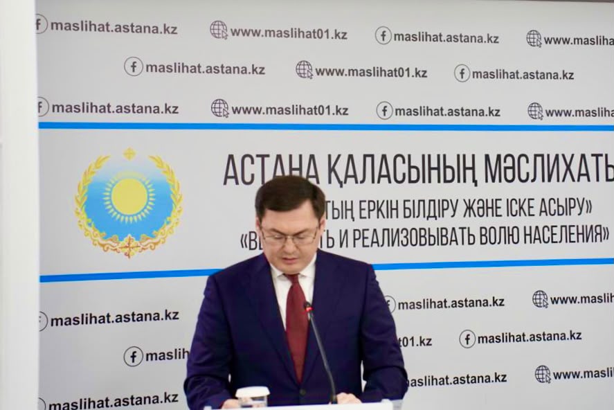 Астана қаласының мәслихатының алдында сөйлеп тұрған костюм киген ер адам.