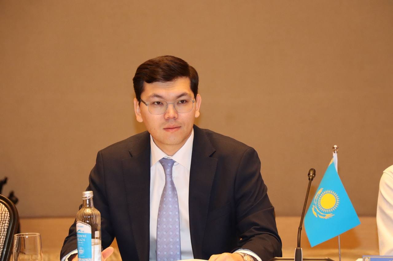 Костюм киген, көзілдірік таққан ер адамның портреті. Жанында Қазақстан Республикасының шағын туы тұр.