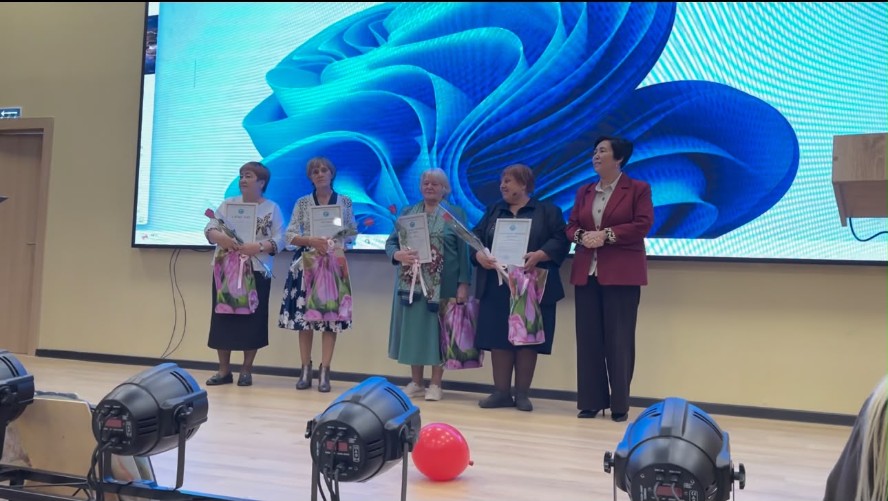 Сахнада грамоталары мен гүлдері бар төрт әйел және олардың жанында тұрған жүргізуші.