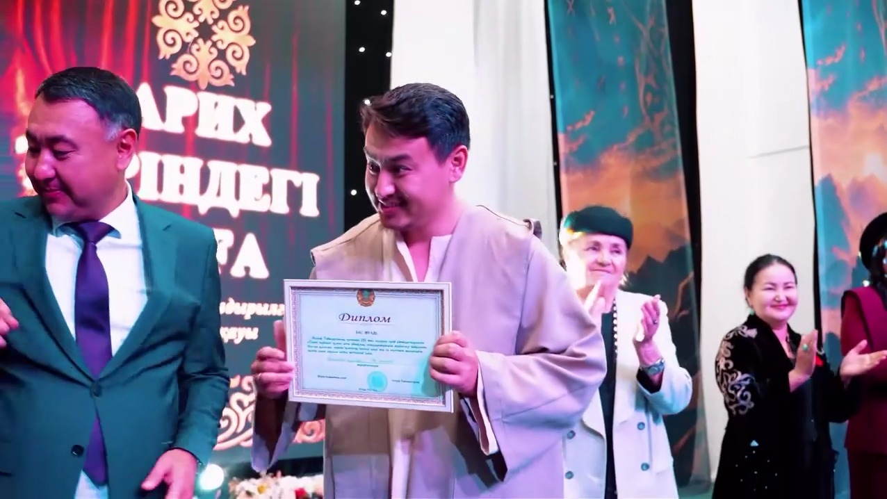 Диплом алған ұлттық киімдегі ер адам және оны қолдап тұрған адамдар.