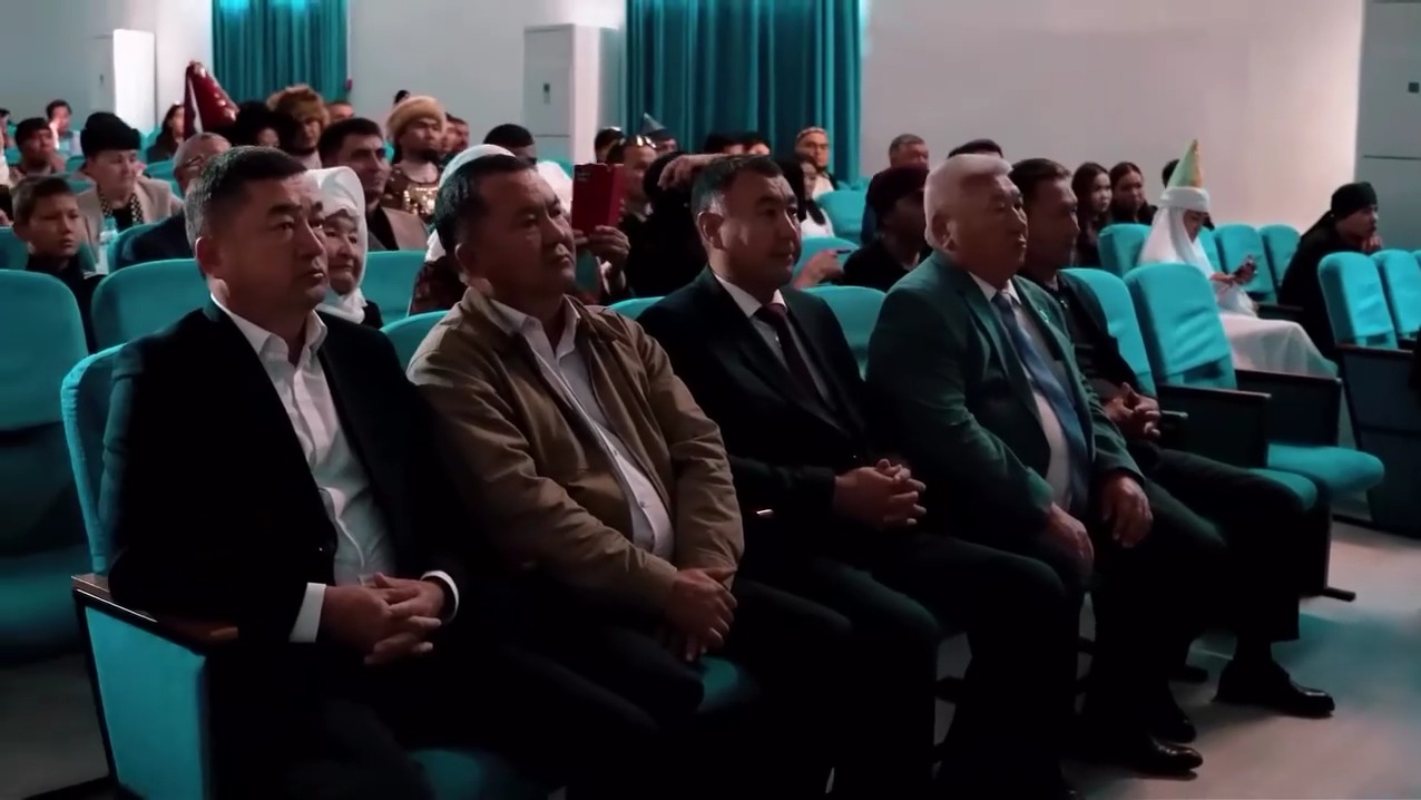 Жиынға жиналған, көк орындықтарда отырған ерлер мен әйелдер тобы.