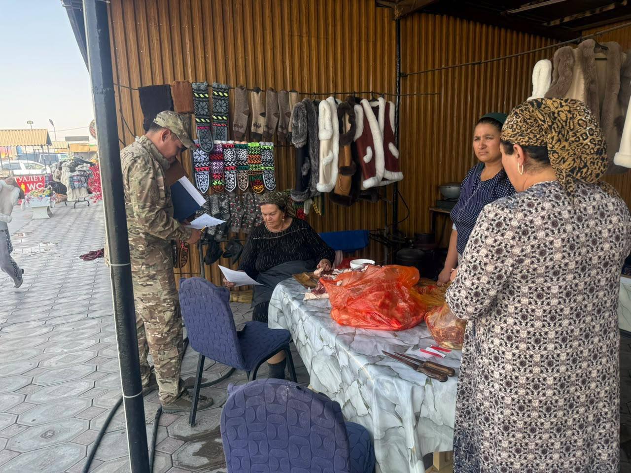 Базардағы сауда нүктесінде құжаттарды тексеріп жатқан әскери қызметші және сатушы әйелдер.