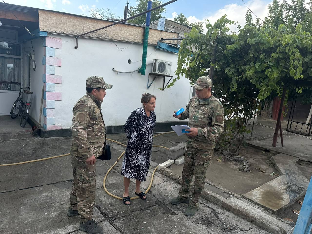 Әскери формадағы екі адам мен әйелдің үйдің ауласында сөйлесіп тұрғаны.