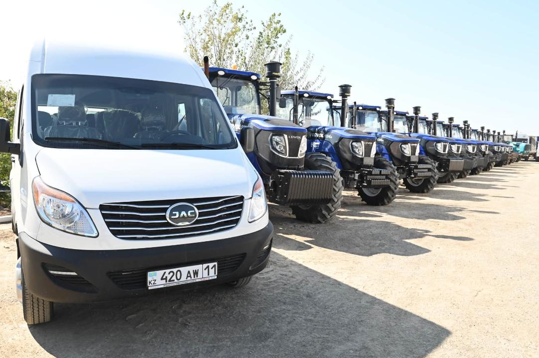 Ақ түсті JAC микроавтобусы және оның жанында тізіліп тұрған көк түсті тракторлар.