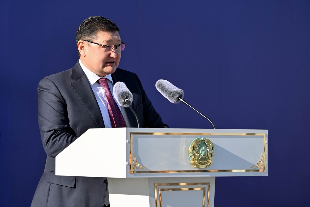 Көк фон алдындағы трибунада сөйлеп тұрған костюм киген ер адам.