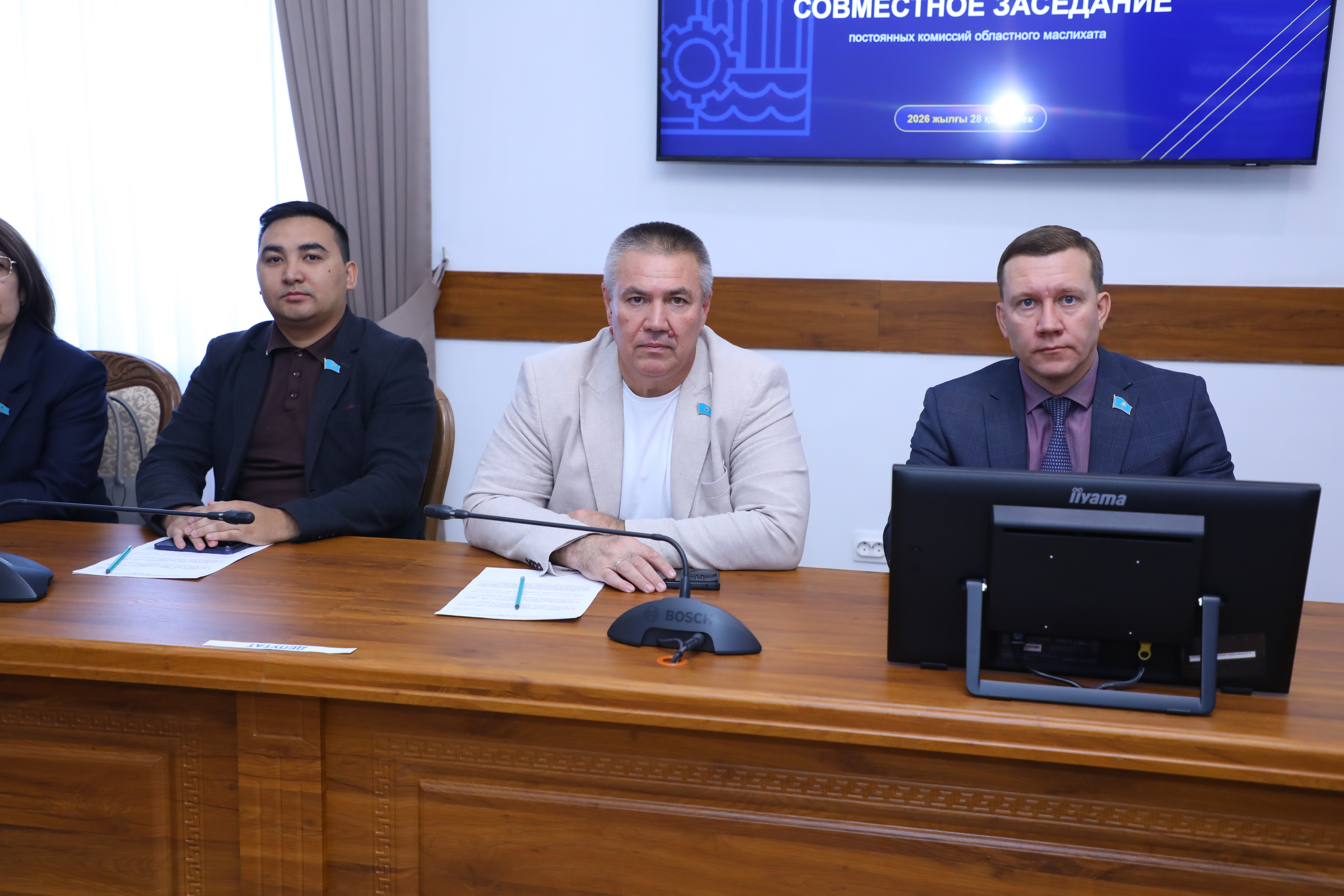 Үстел басында отырған үш ер адам және артқы жақтағы ақпараттық экран.