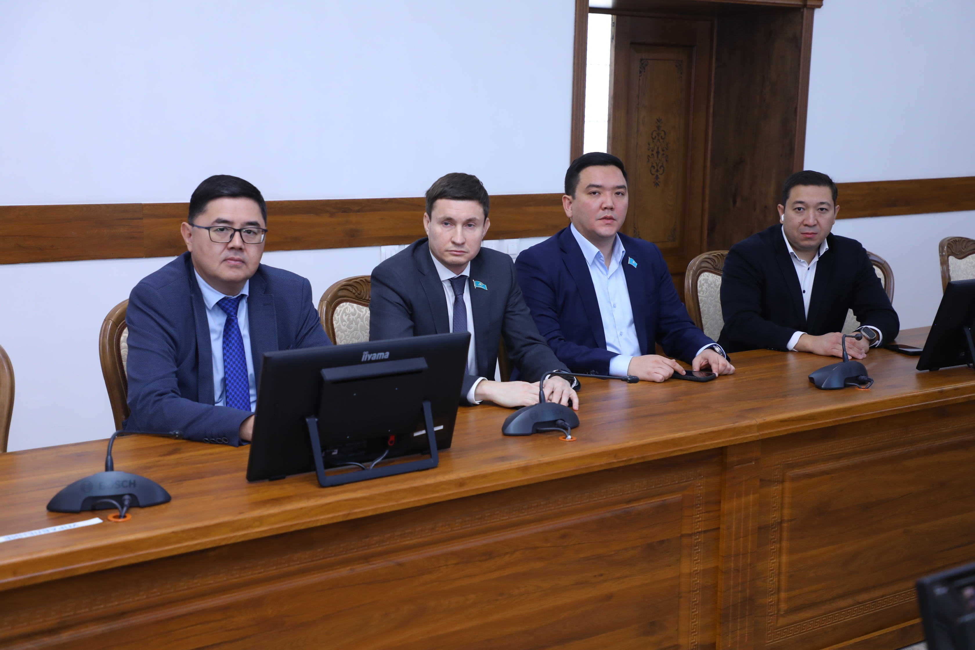 Жиналыс үстелінде отырған костюм киген төрт ер адам.