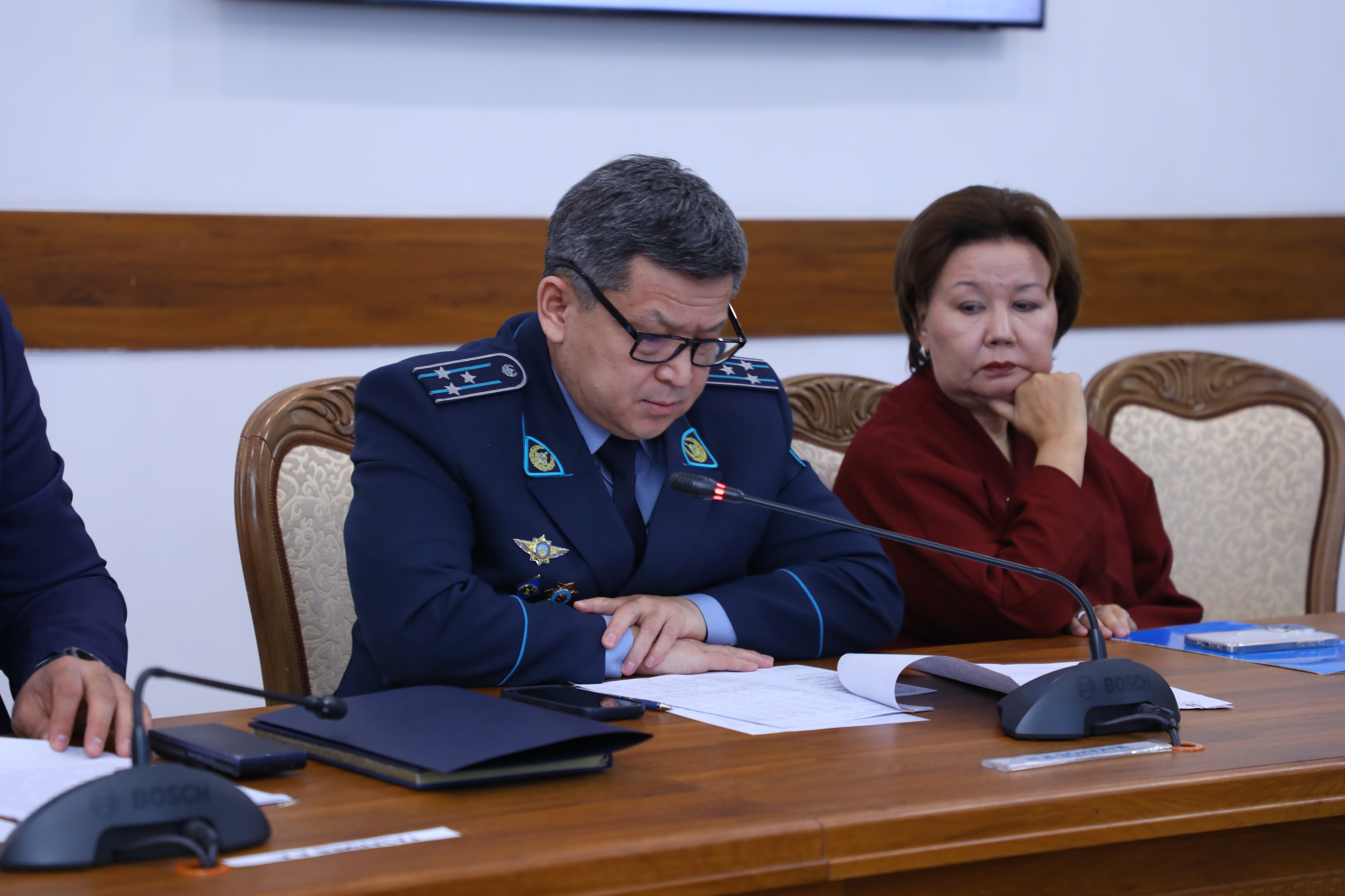 Форма киген ер адам мен қызыл киімді әйел жиналыста отыр.