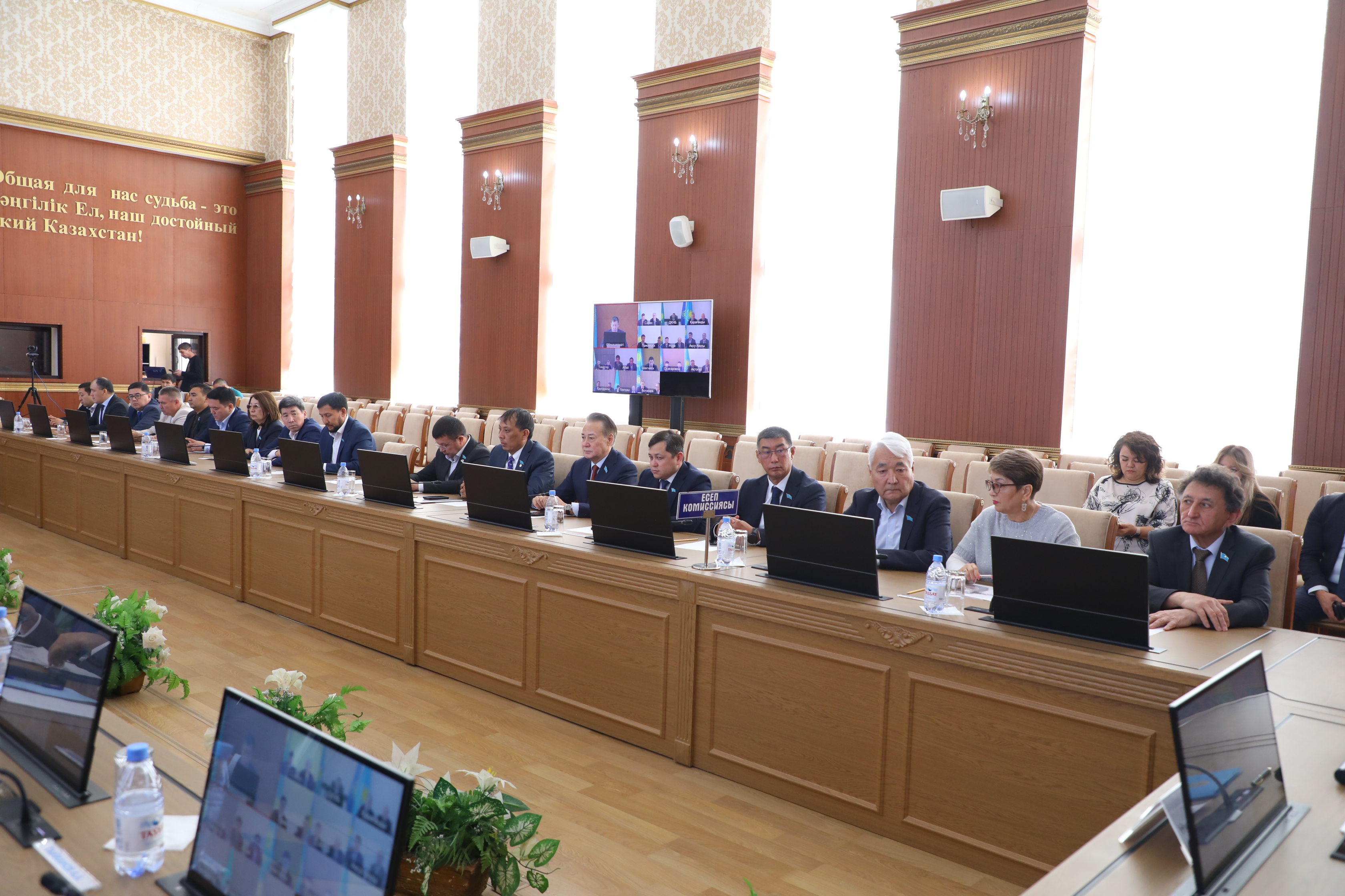Жиналыс залында ноутбуктермен отырған ресми адамдар тобы.