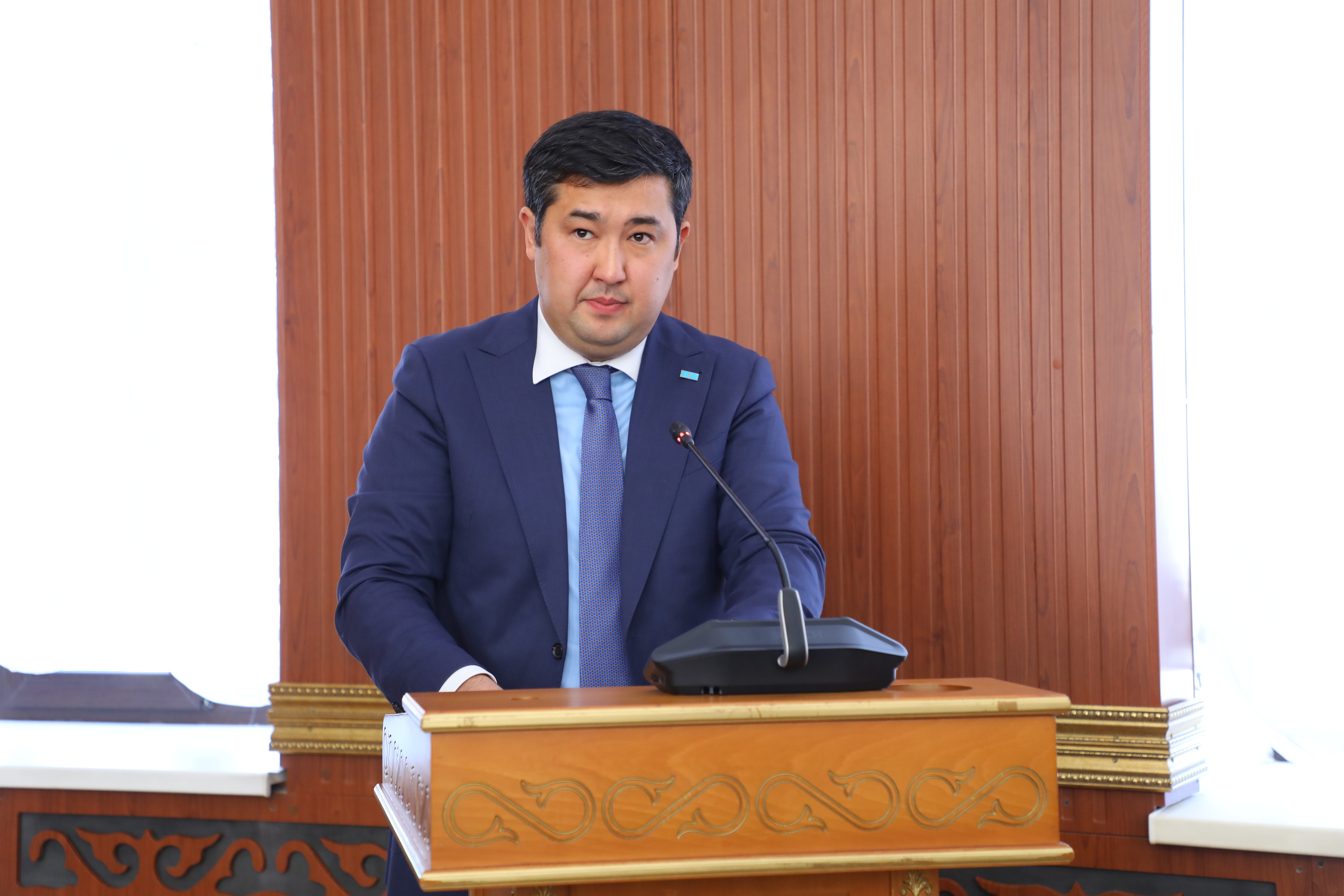 Көк костюм киген ер адам трибунада сөйлеп тұр.