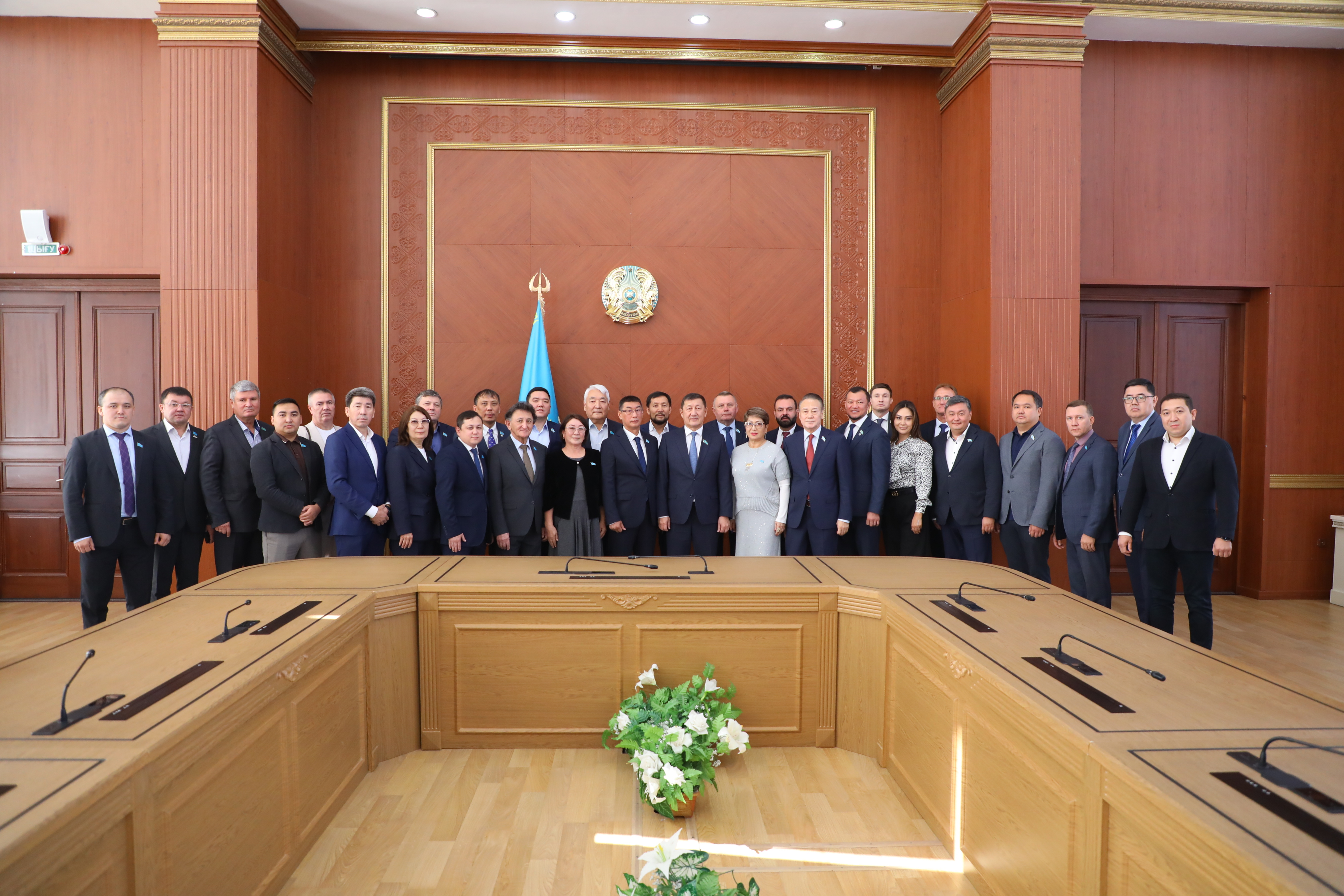 Ресми киімдегі адамдар тобы Қазақстан Республикасының мемлекеттік рәміздері тұрған залдың ішіндегі топтық суреті.