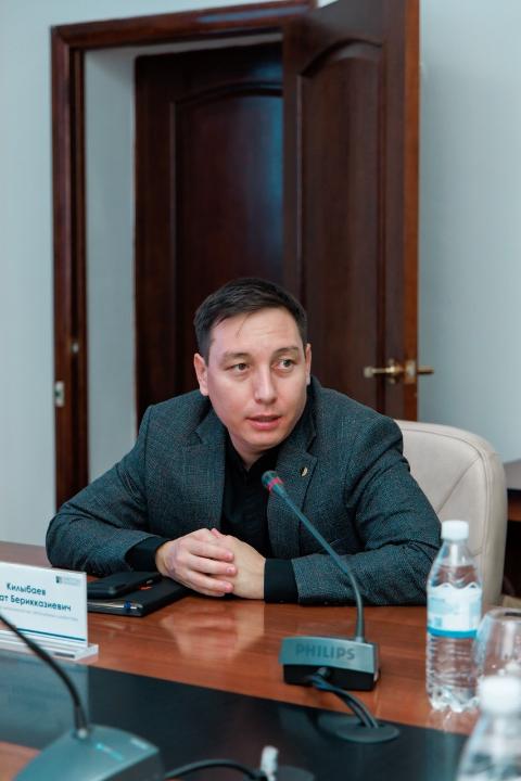Мужчина в сером пиджаке сидит за столом на совещании перед микрофоном.