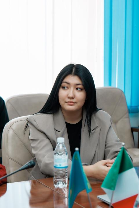 Женщина в бежевом пиджаке сидит за столом на официальном мероприятии.