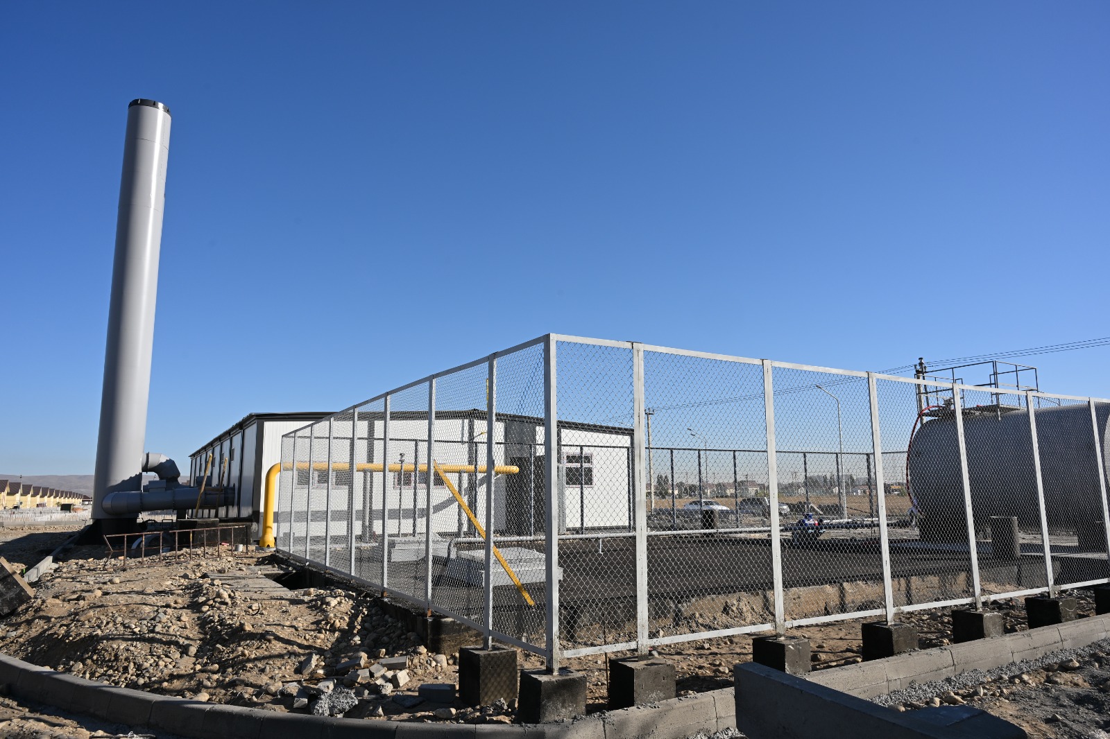 Қоршаулы аумақтағы газ құбырлары, резервуар және модульдік ғимарат.