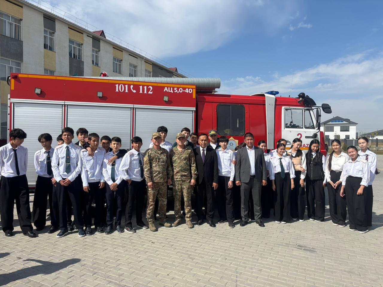 Өрт сөндіру көлігінің алдында тұрған мектеп оқушылары мен мамандардың топтық суреті.