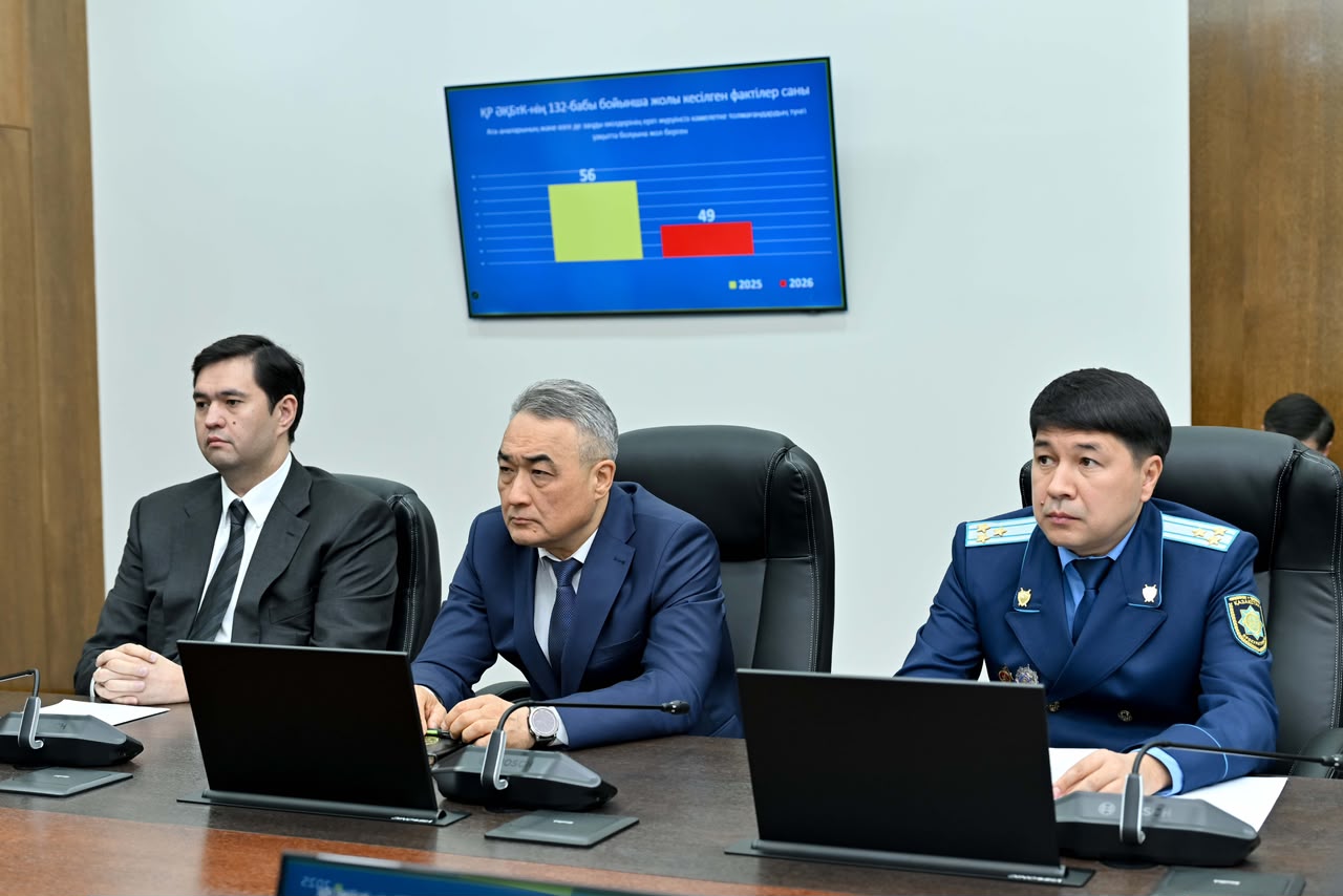 Жиналыста отырған үш ер адам және артқы жақтағы статистикалық мәліметтері бар экран.