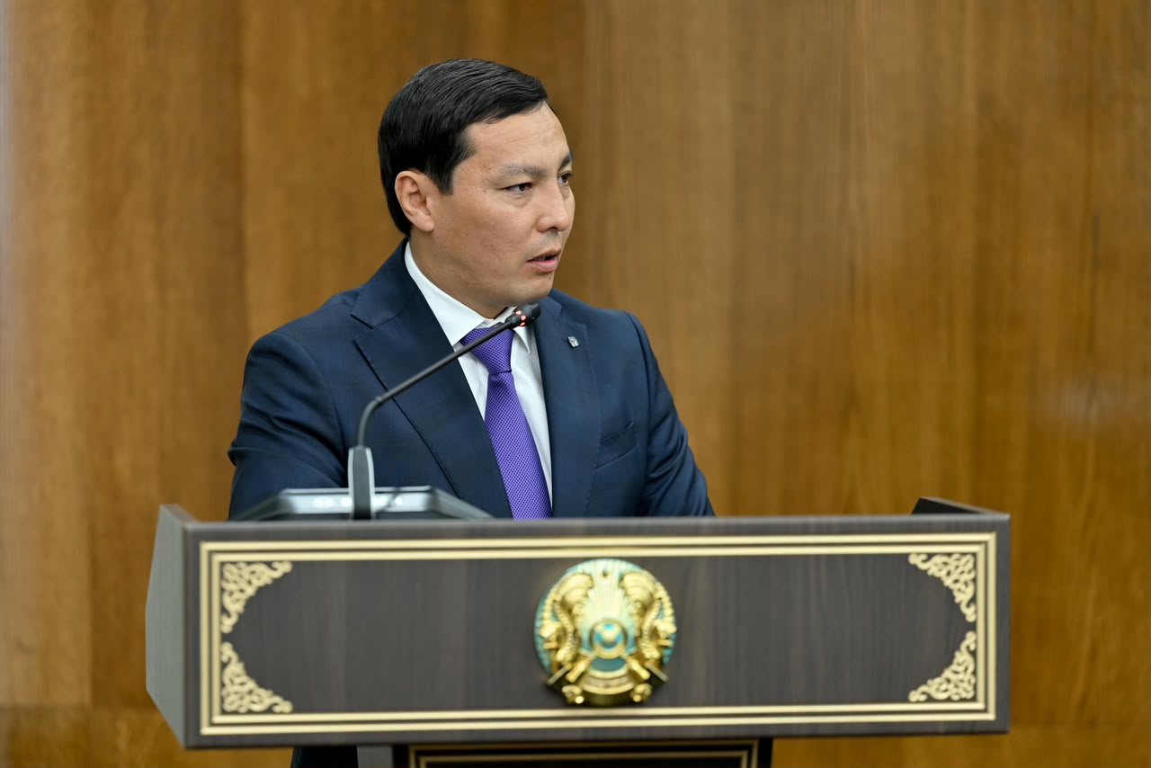 Көк костюм мен күлгін галстук киген ер адам трибунада сөйлеп тұр.