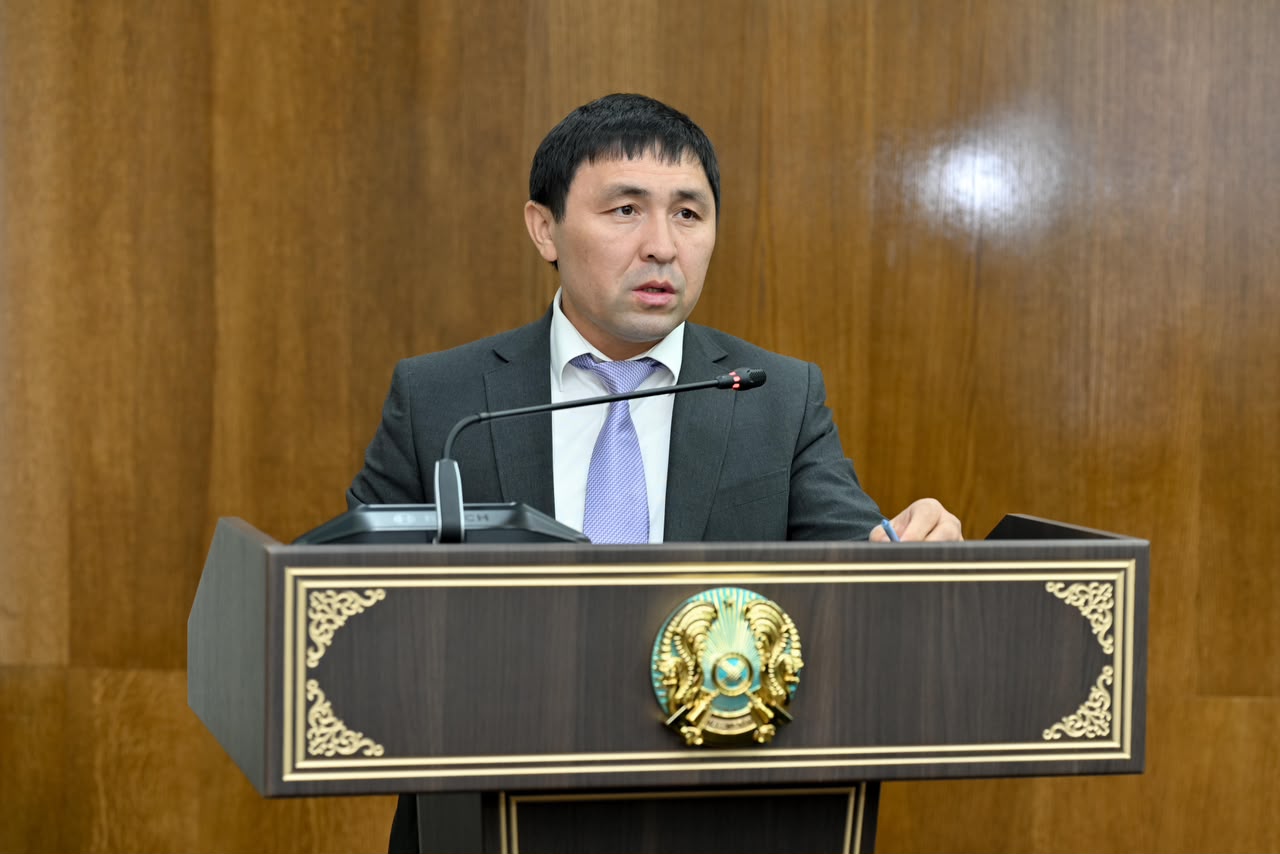 Қазақстан Республикасының мемлекеттік рәмізі бар трибунада сөйлеп тұрған костюм киген ер адам.