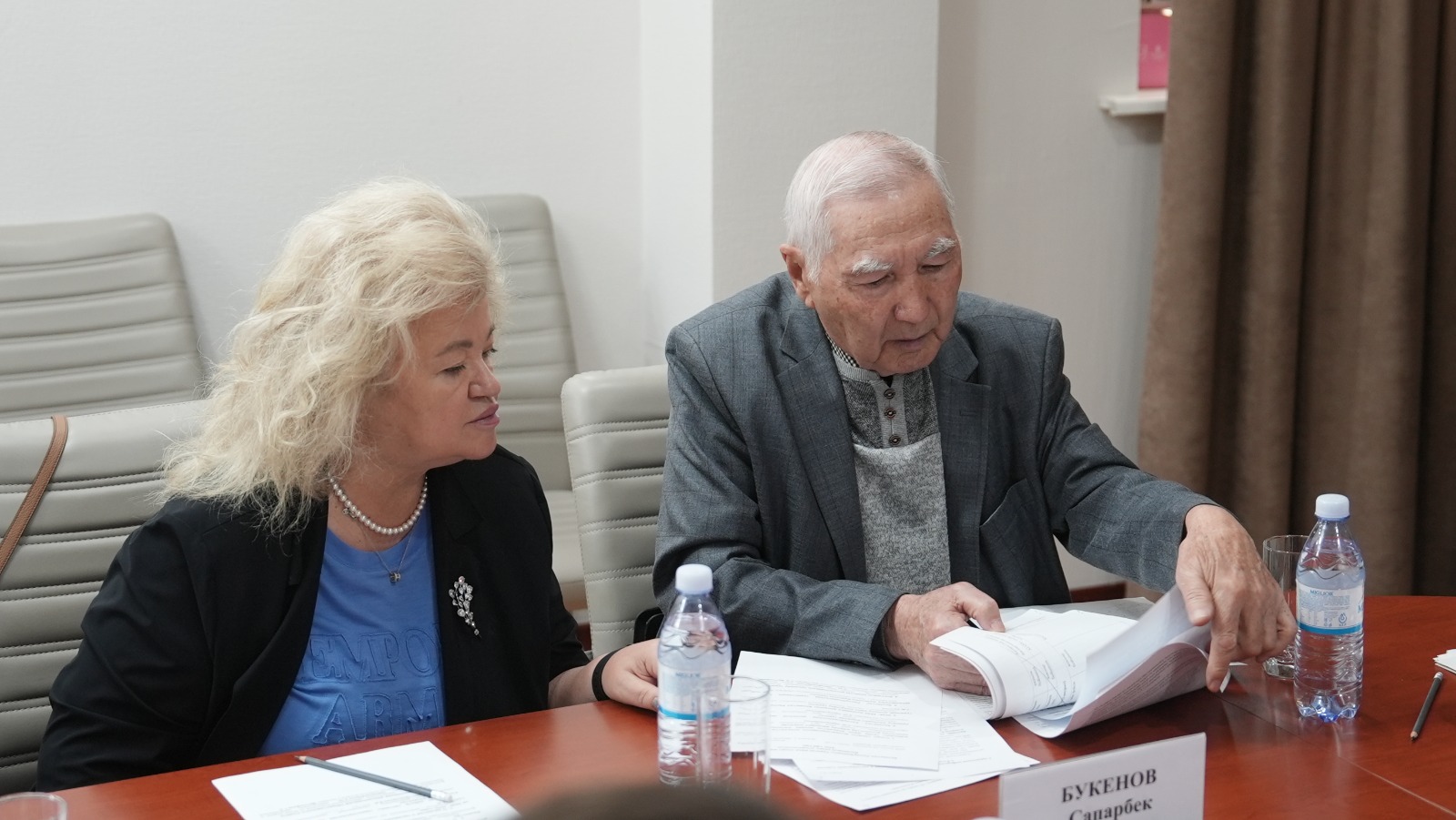 Келіссөз үстелінде құжаттарды қарап отырған екі адам.