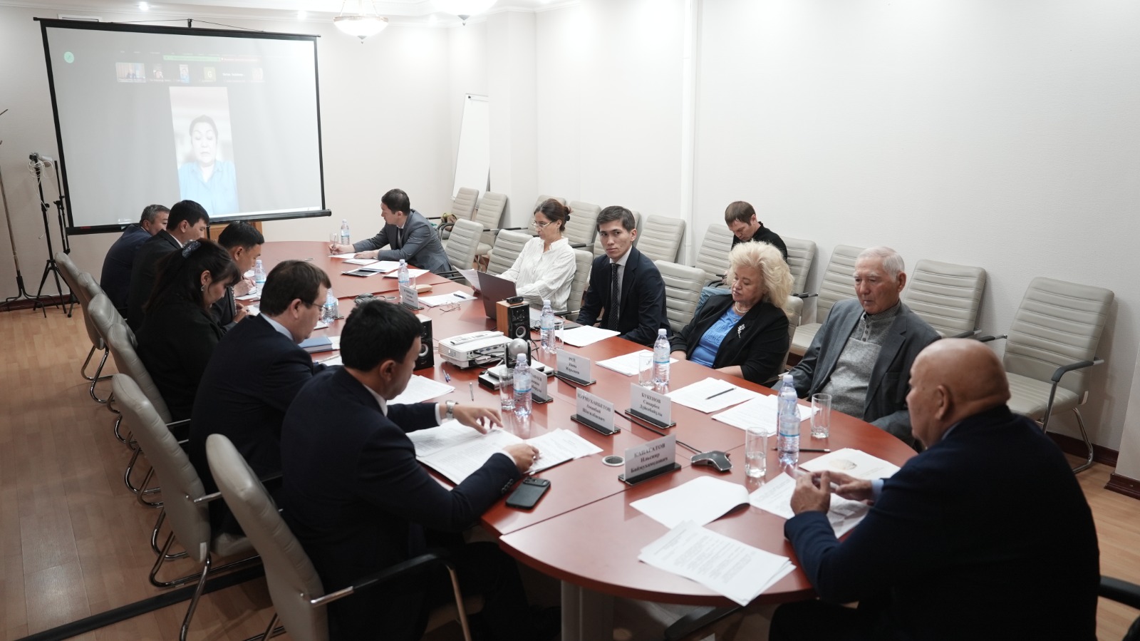 Жиналыс залында үстел басында отырған адамдар және экрандағы бейнебайланыс.