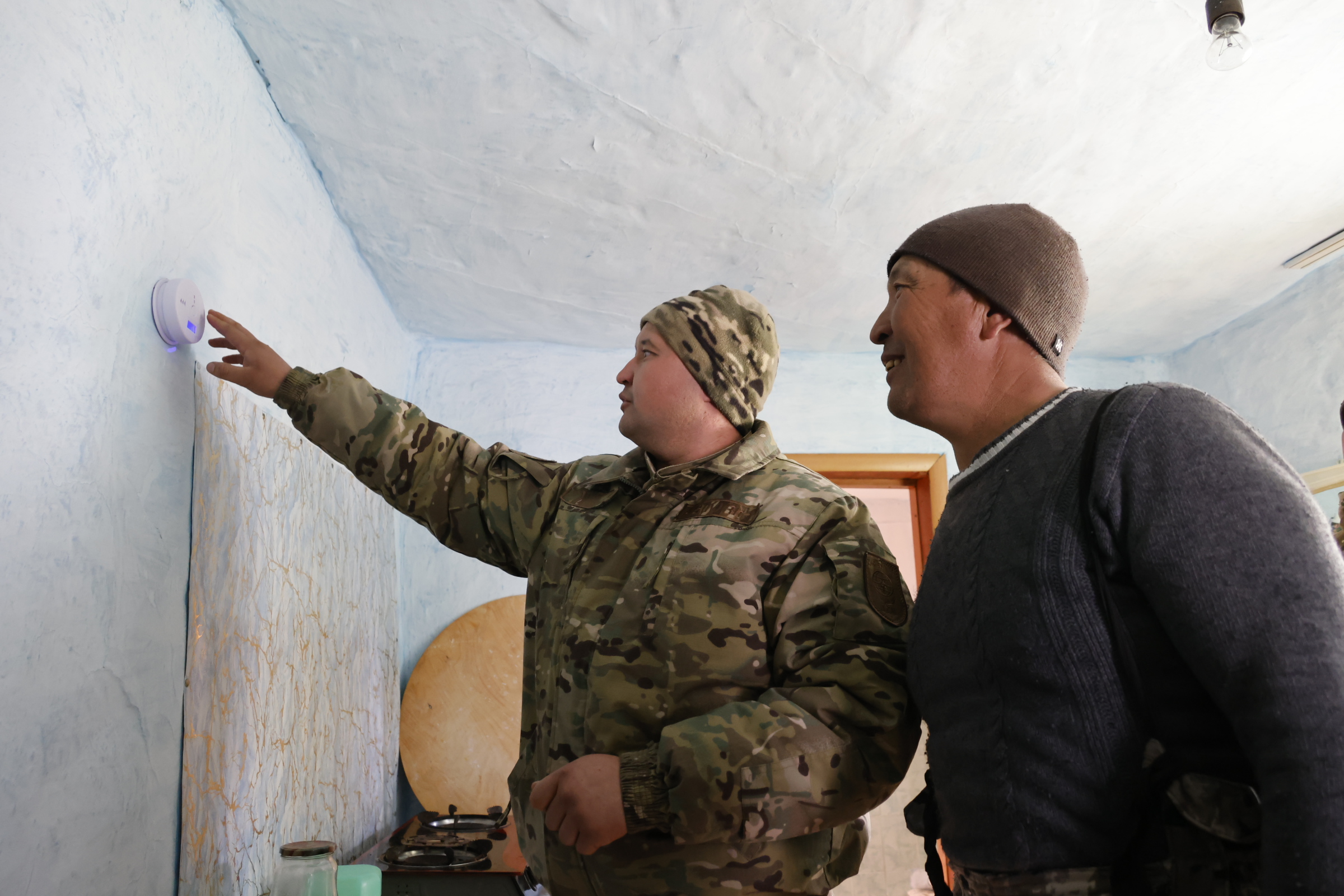 Сотрудник ДЧС показывает мужчине в шапке датчик, закрепленный на голубой стене в помещении.