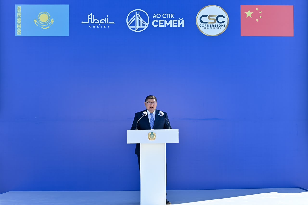 Көк фонды және Қазақстан мен Қытайдың тулары, компания логотиптері орналасқан трибунада сөйлеп тұрған ер адам.