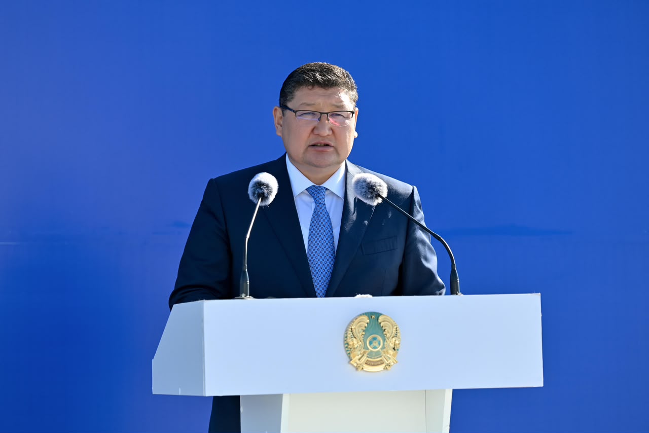 Көк фон алдында, Қазақстан Республикасының елтаңбасы бар трибунада сөйлеп тұрған костюм киген ер адам.