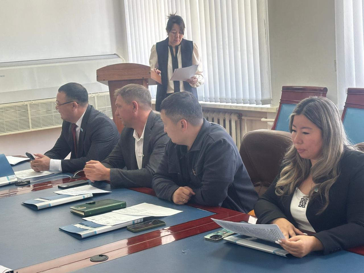 Жиналыс кезінде үстел басында отырған төрт адам және баяндама жасап тұрған әйел.