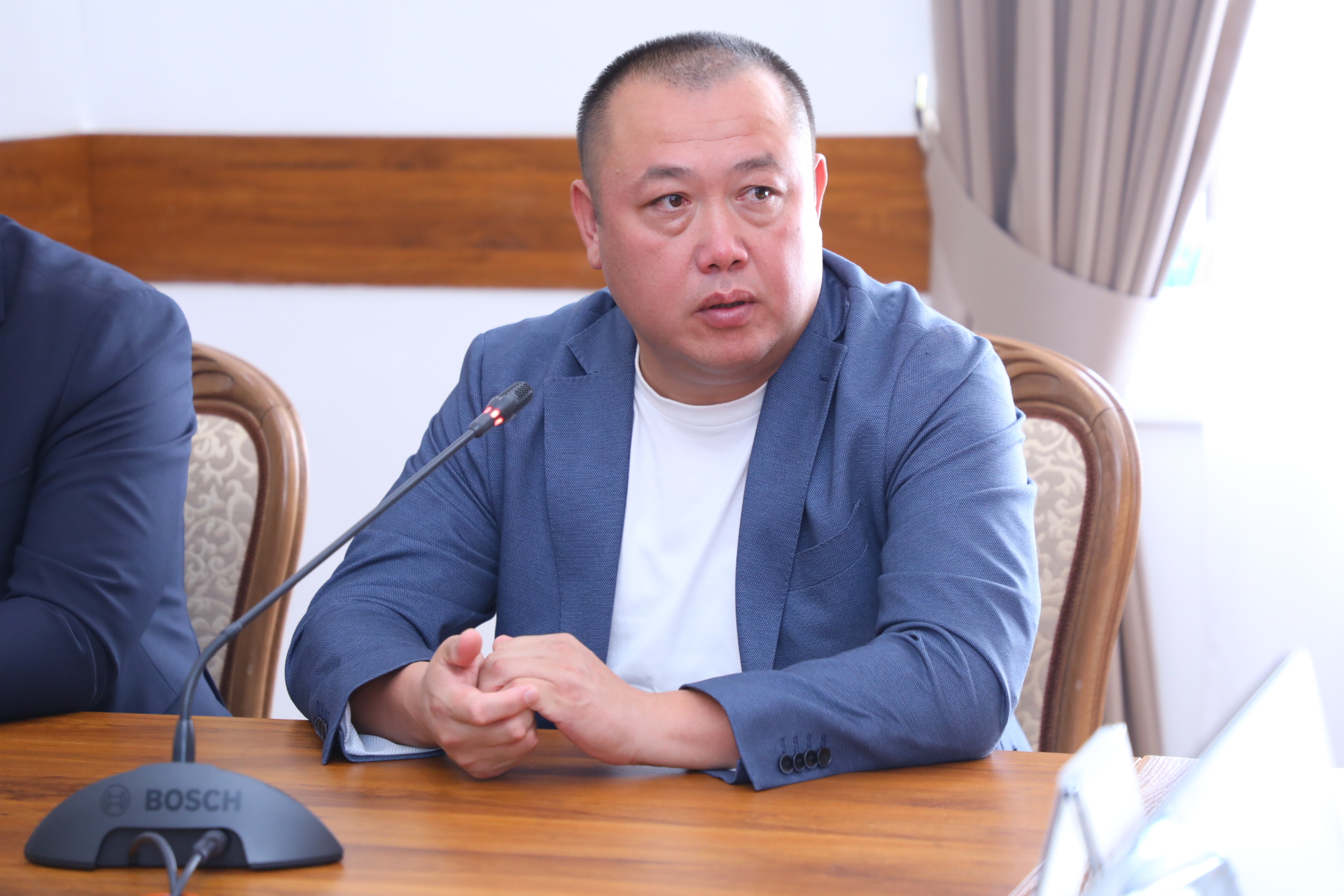 Көк пиджак киген ер адам микрофонның алдында отырып сөйлеп тұр.