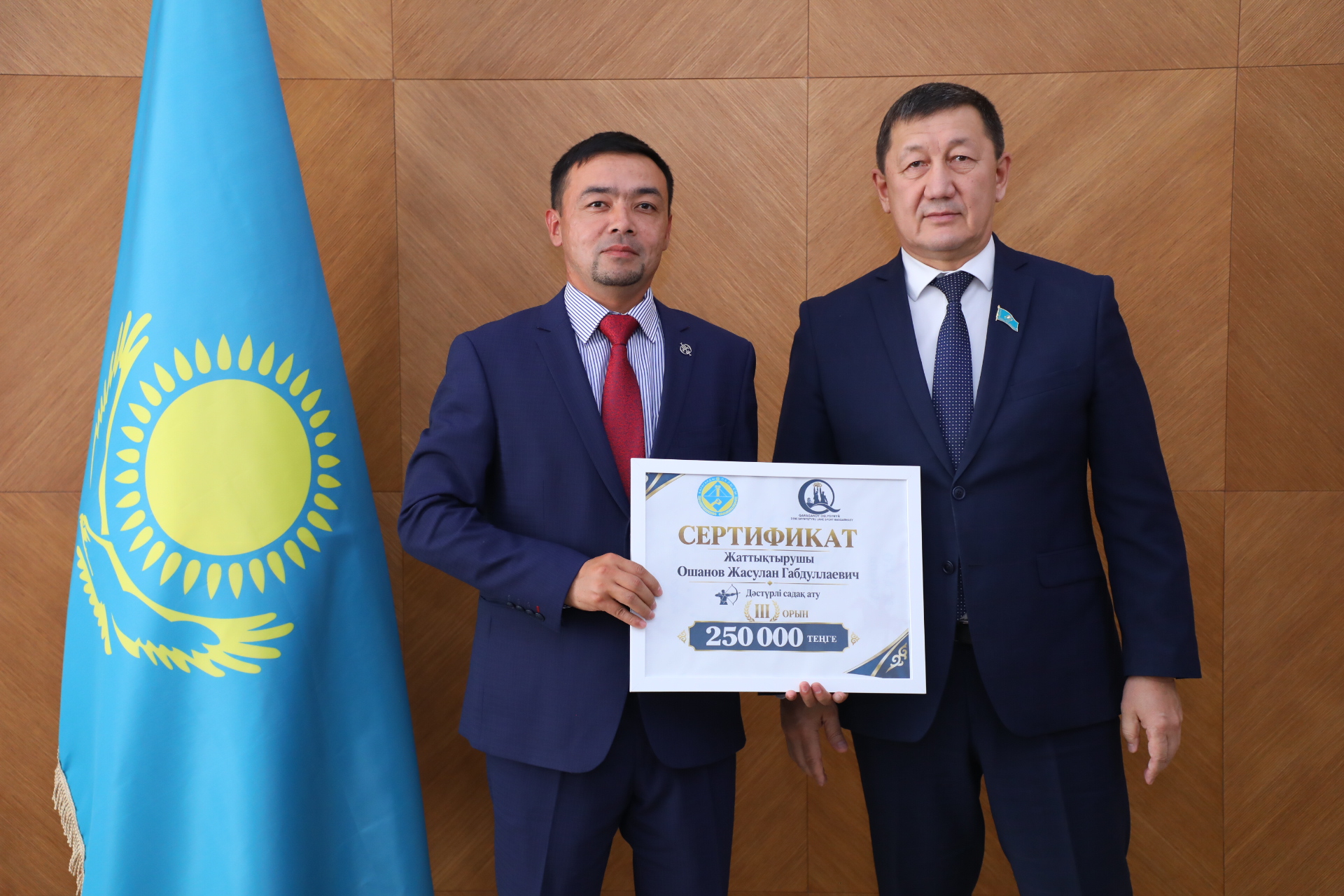 Қазақстан туының жанында екі ер адам сертификат ұстап тұр.