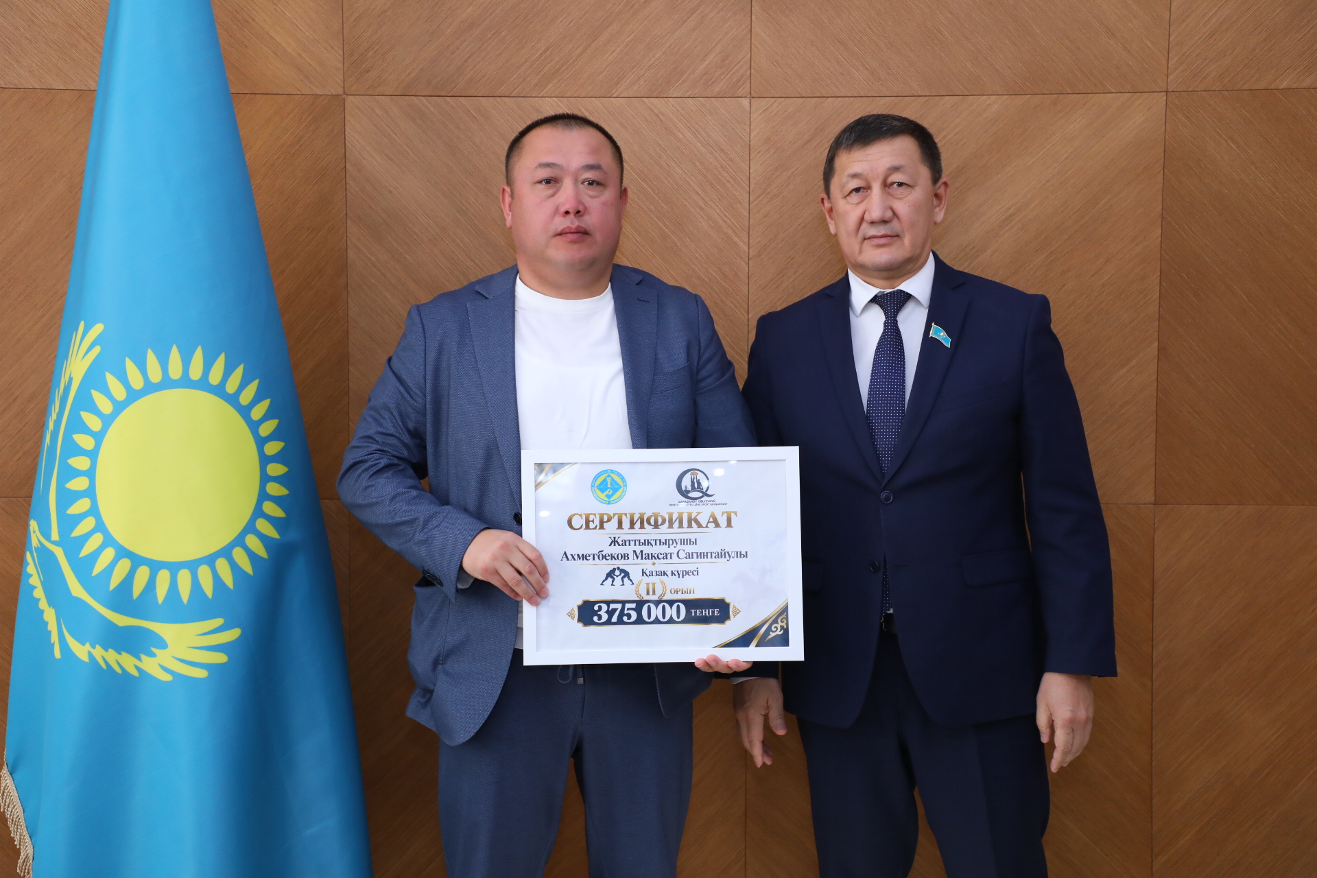 Қазақстан туының жанында екі ер адамның сертификат ұстап тұрған суреті.