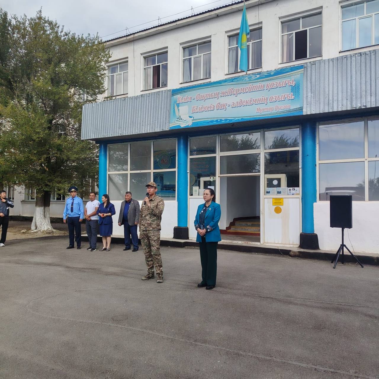 Мектептің алдында жиналған әскери және азаматтық киімдегі адамдар тобы.