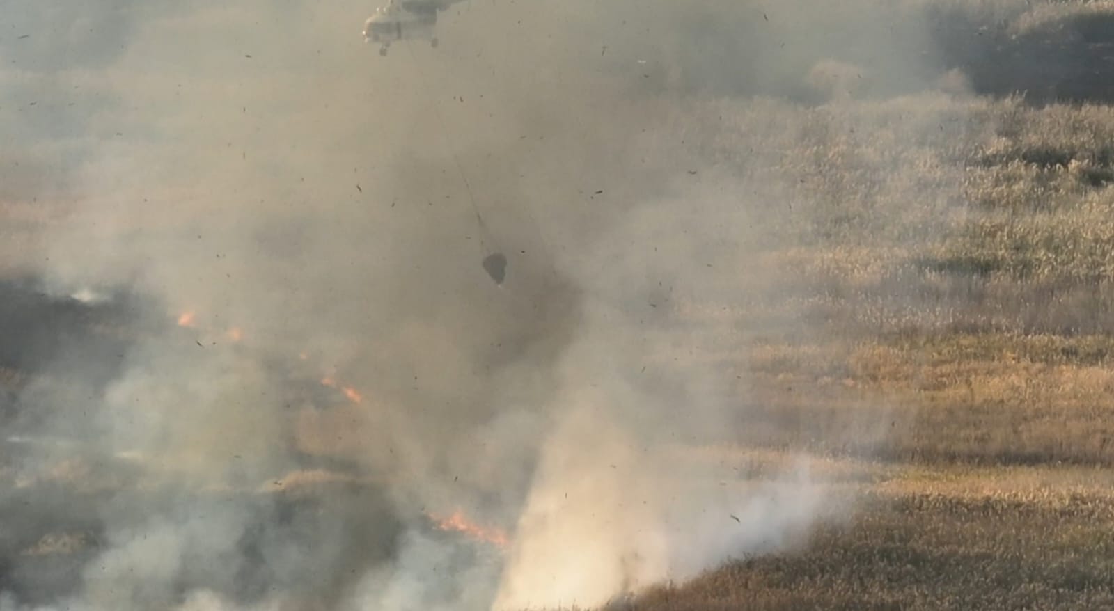 Вертолет сбрасывает воду на пожар в поле, окутанное густым дымом.