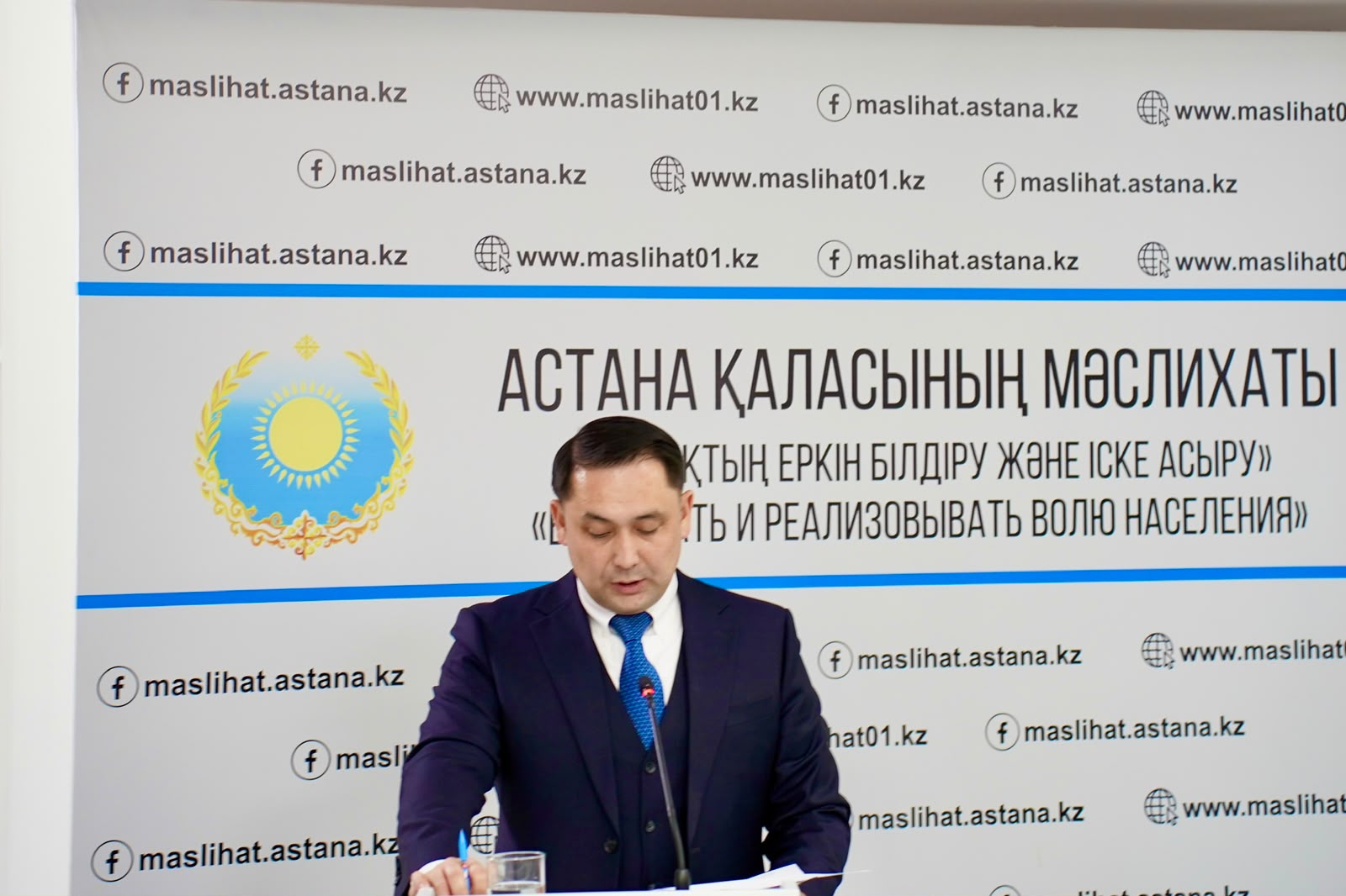 Мужчина в синем костюме выступает на фоне баннера Маслихата города Астаны с государственным гербом Казахстана.