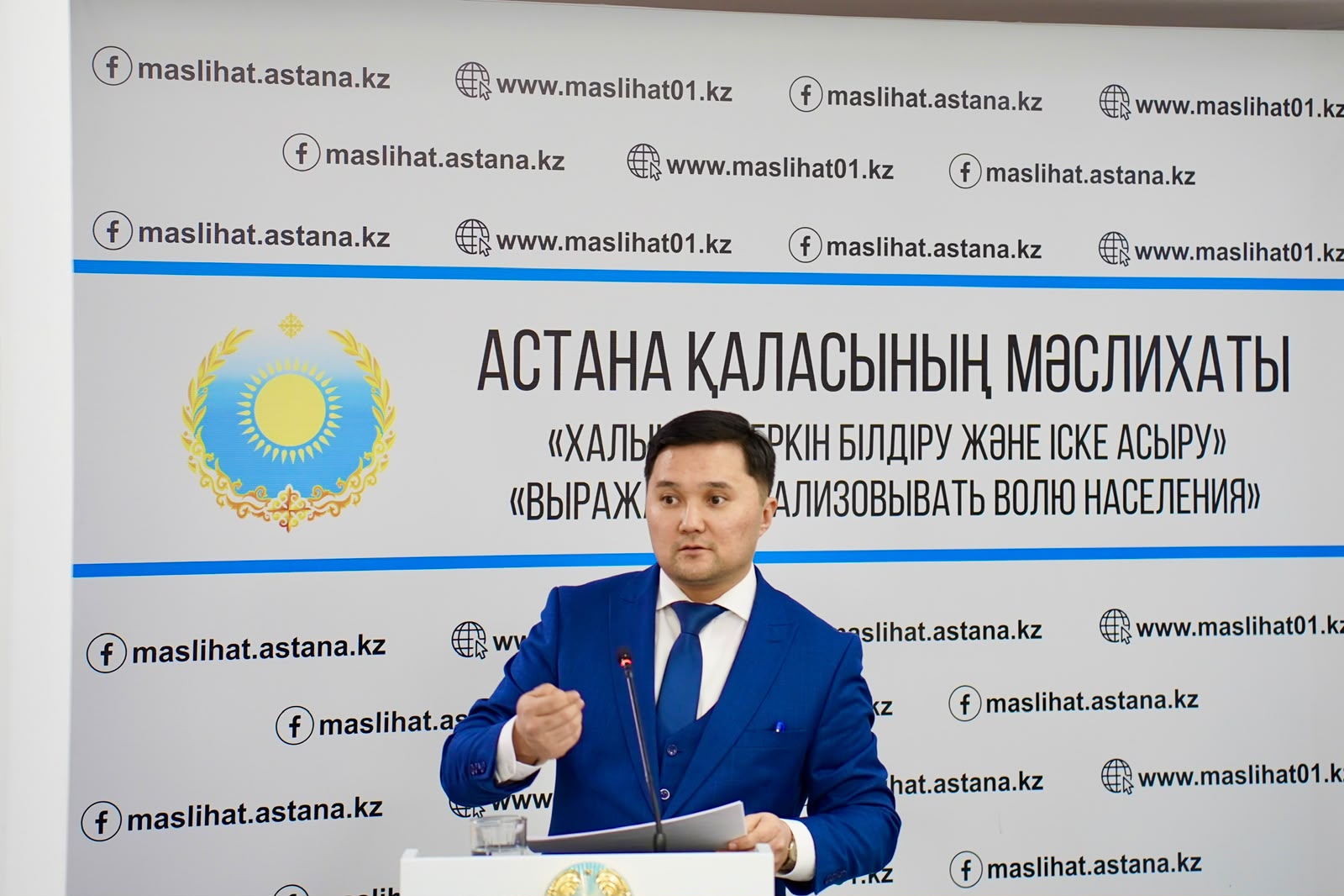 Мужчина в синем костюме выступает на фоне баннера Маслихата города Астаны.