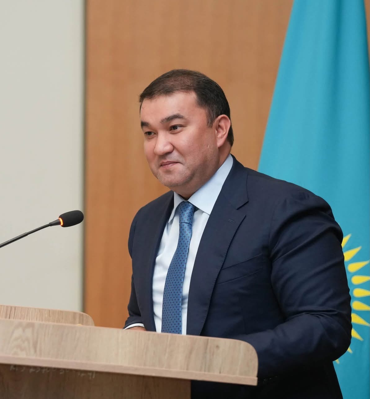 Қазақстан туының алдында трибунада тұрған костюм киген ер адам.