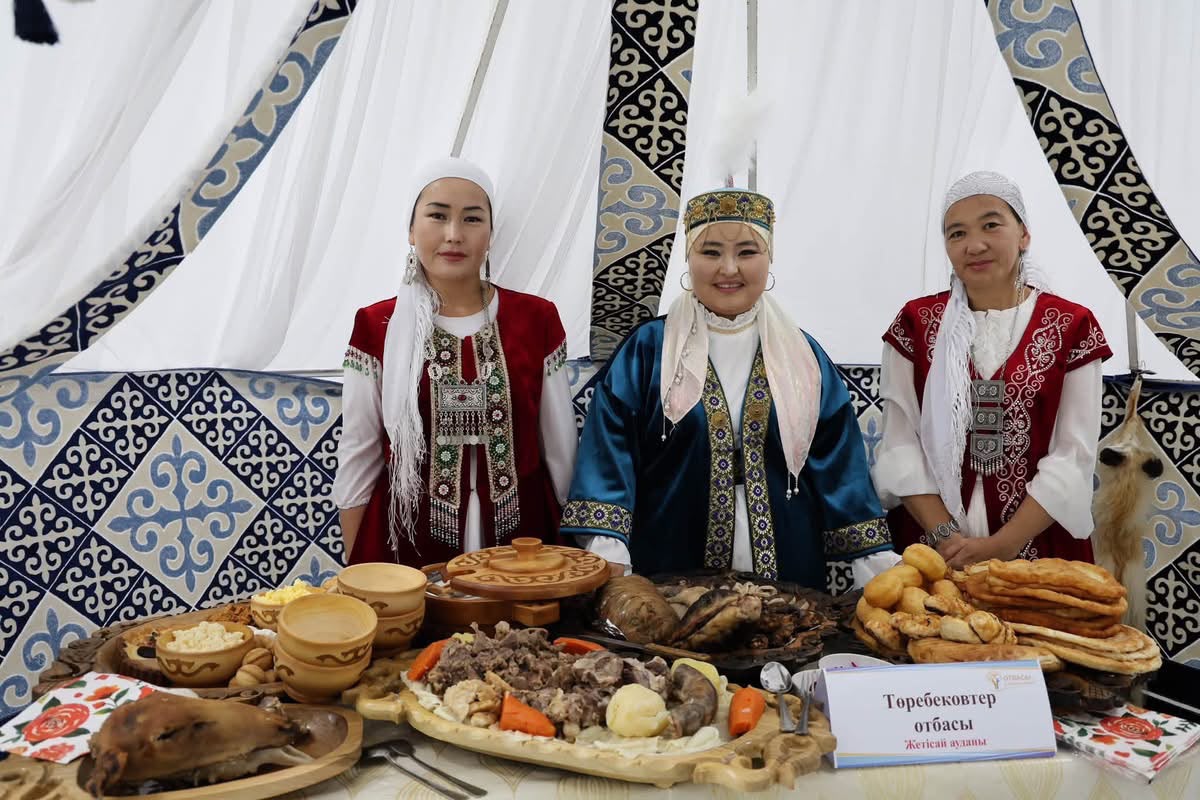 Ұлттық киім киген үш әйел киіз үй ішіндегі қазақша дастархан жанында тұр.