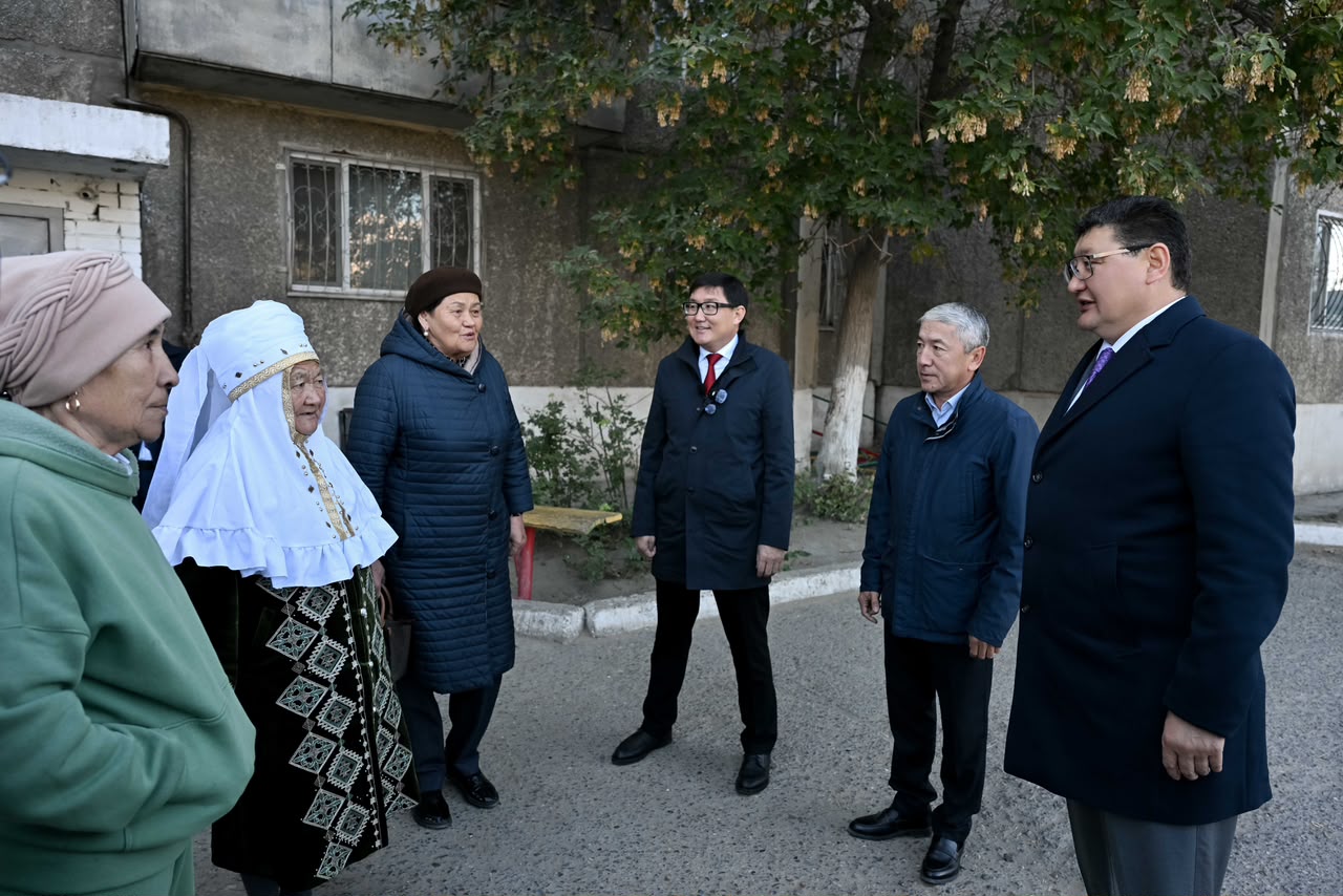 Көшедегі үйдің алдында тұрып, сөйлесіп жатқан қариялар мен костюм киген ер адамдар.