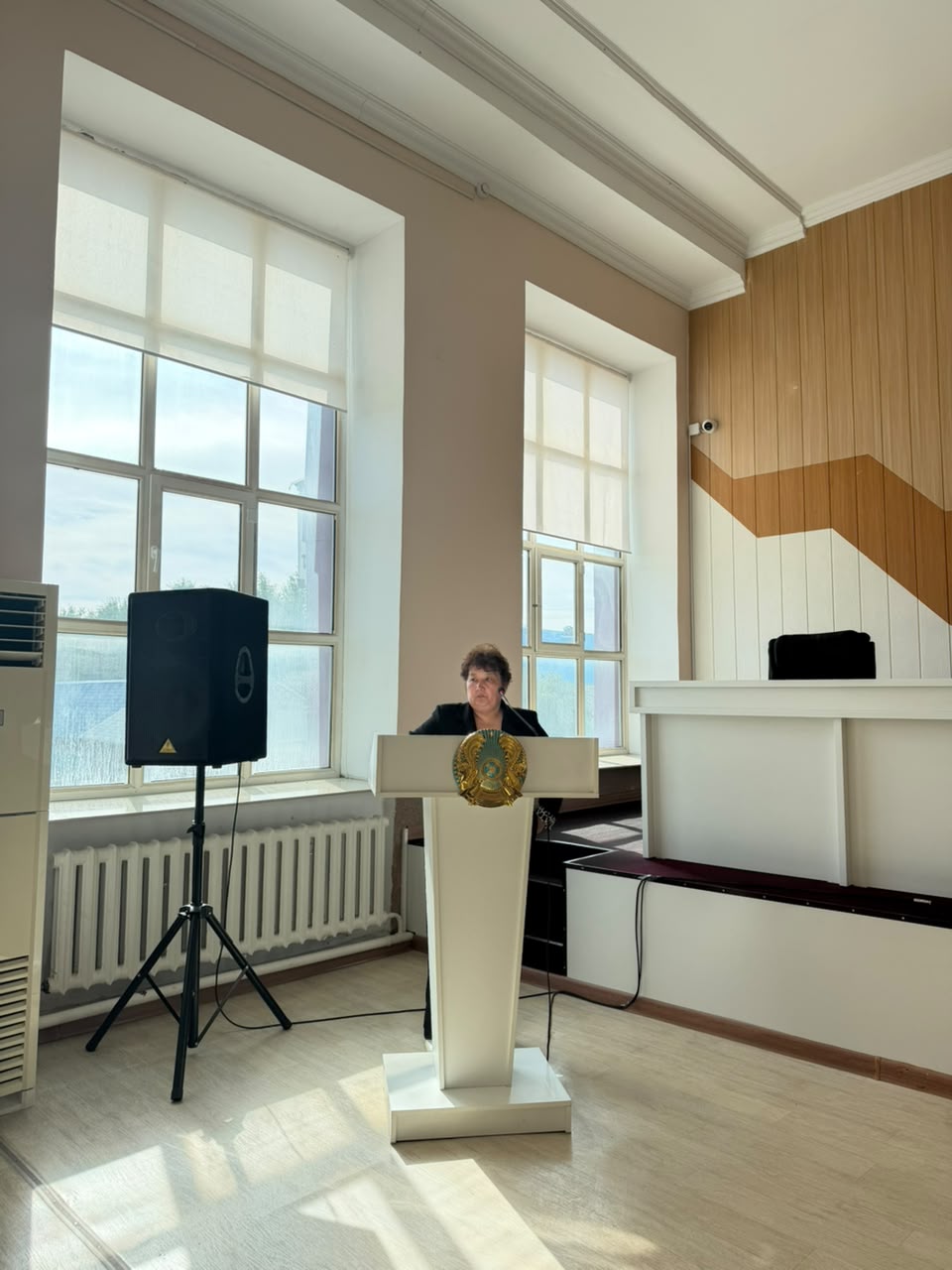 Қазақстан Республикасының елтаңбасы бар трибунада сөйлеп тұрған әйел.
