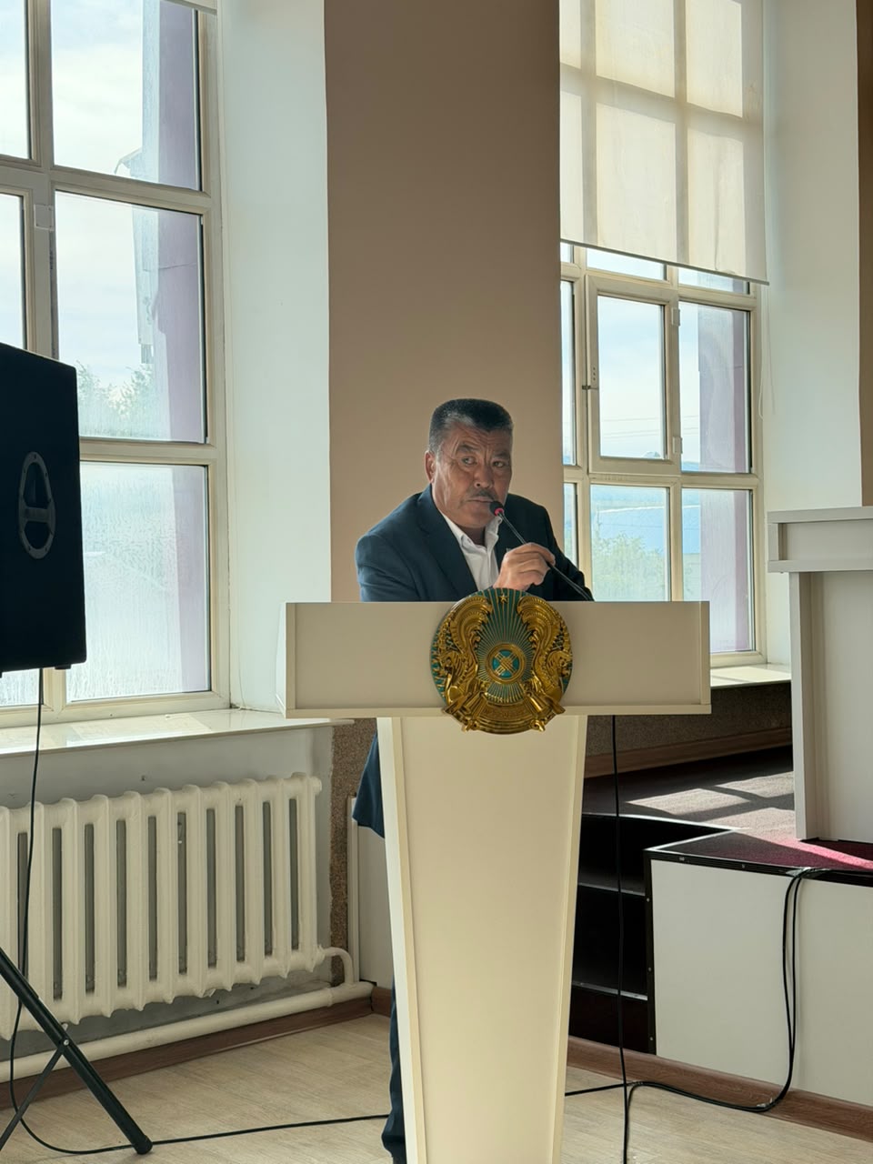 Трибунада микрофонмен сөйлеп тұрған костюм киген ер адам.