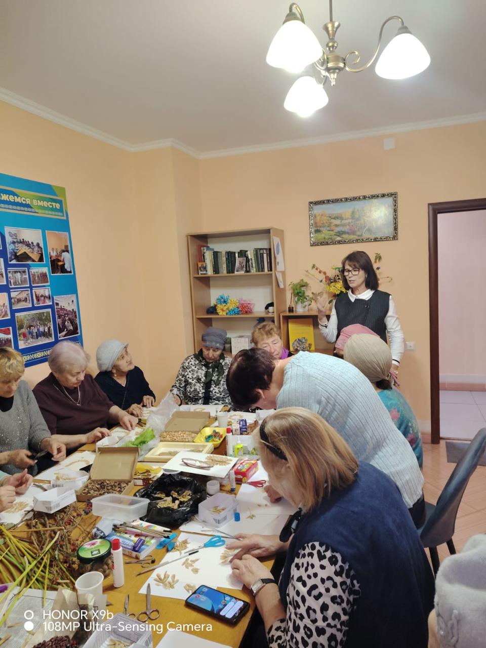 Егде жастағы әйелдер тобы дастархан басында қолөнермен айналысады.