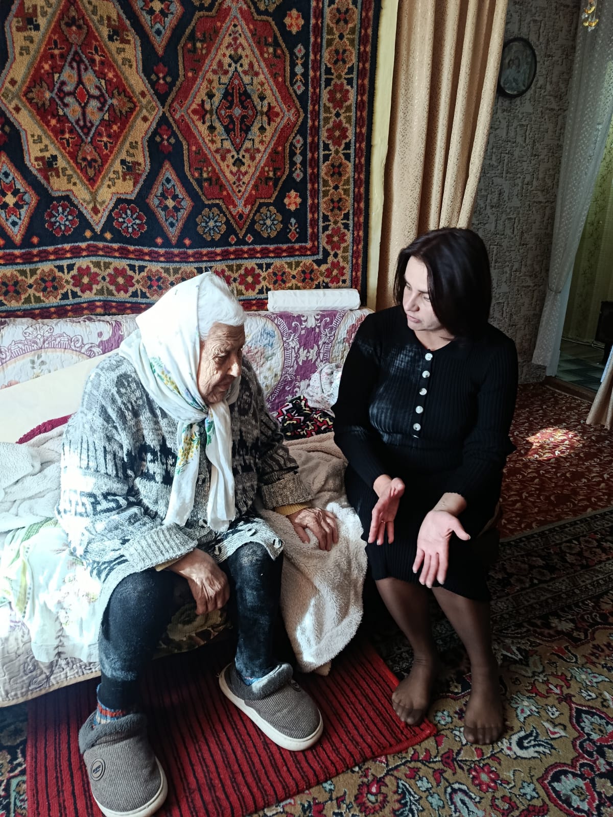 Қара көйлек киген жас әйел кілемдері бар бөлмедегі диванда отырған орамал киген қарт әйелмен сөйлеседі.