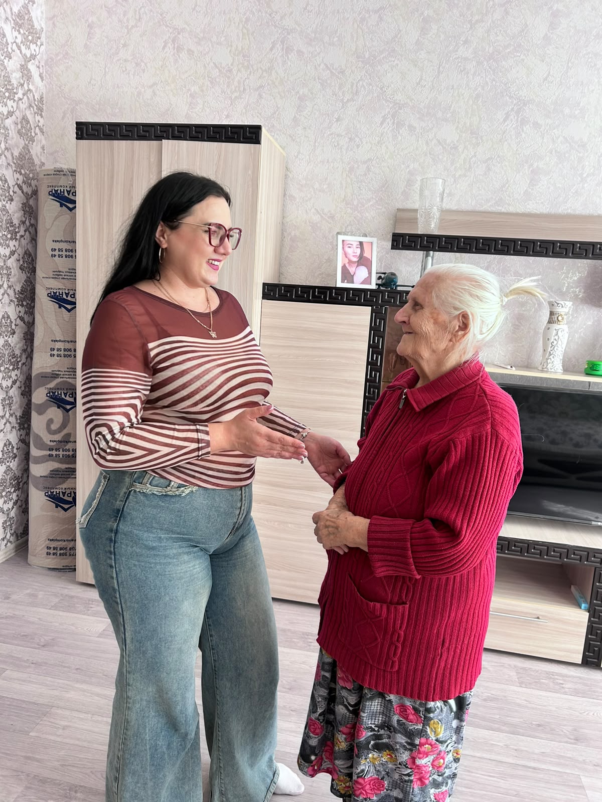 Жолақты үстіңгі және джинсы киген жас әйел бөлменің интерьерінде қызыл кардиган киген қарт әйелмен сөйлеседі.