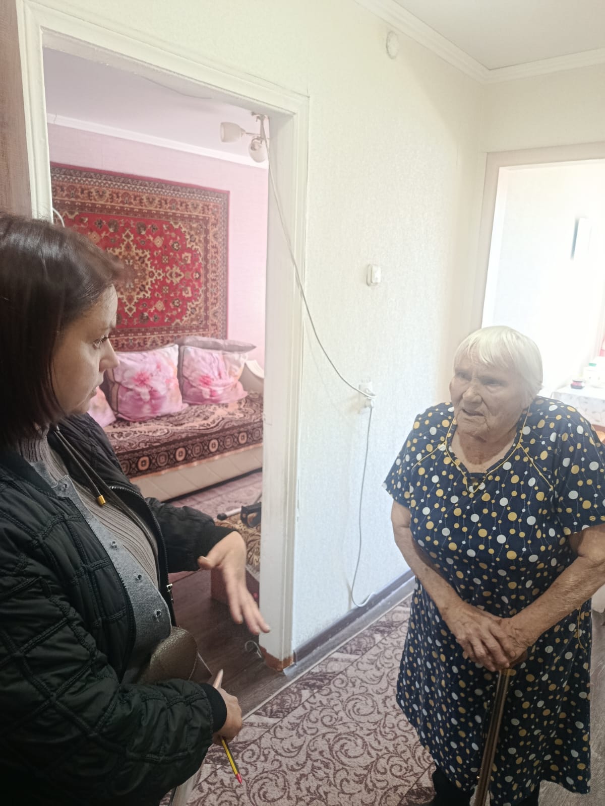 Қара куртка киген жас әйел пәтердің интерьерінде көк полка нүктелі көйлек киген қарт әйелмен сөйлеседі.