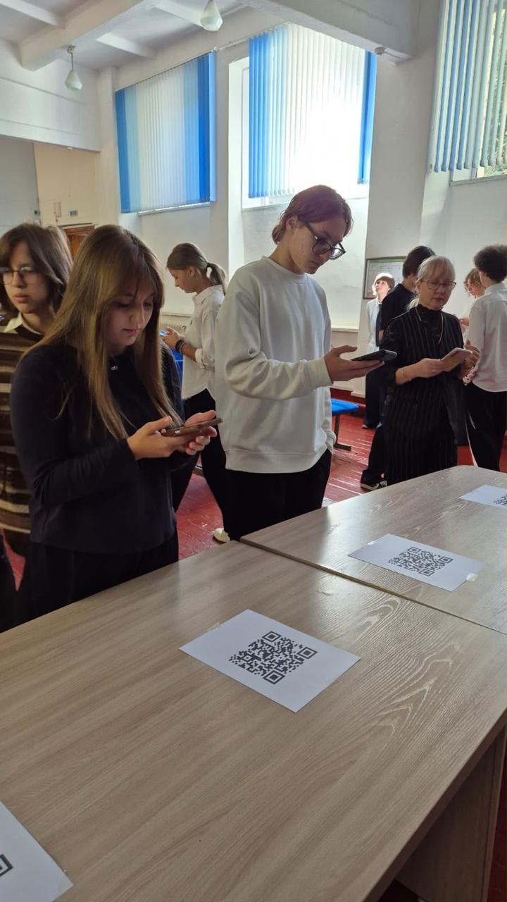 Сыныптағы оқушылар үстел үстіндегі QR-кодтарды смартфондарымен сканерлеп жатыр.