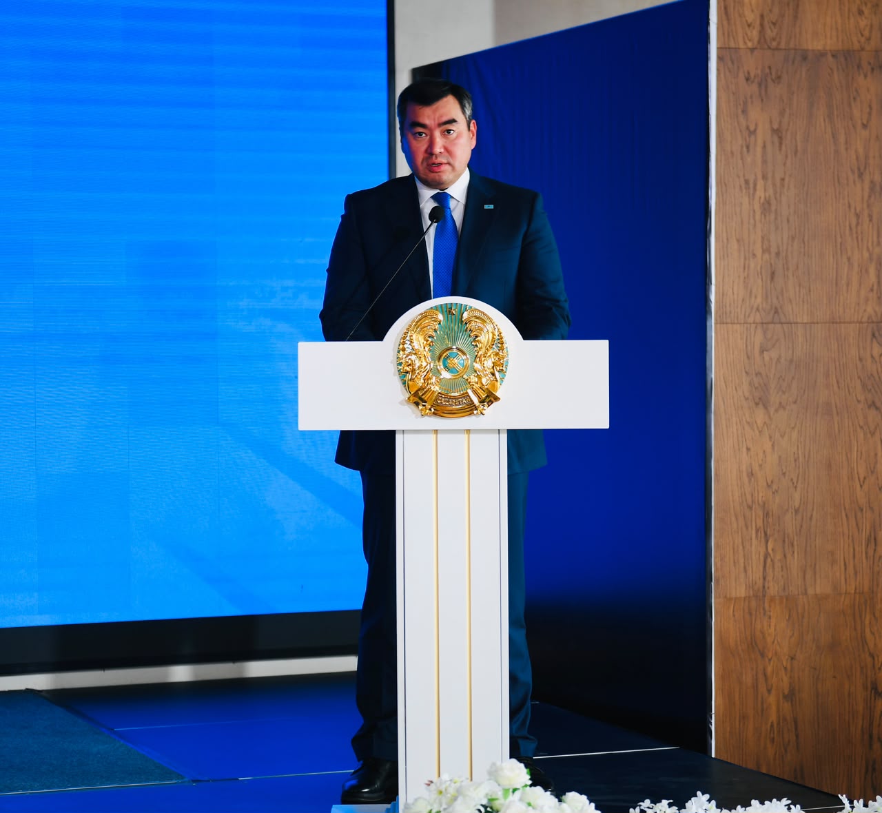 Қазақстан Республикасының мемлекеттік рәмізі бар трибунада сөйлеп тұрған костюм киген ер адам.