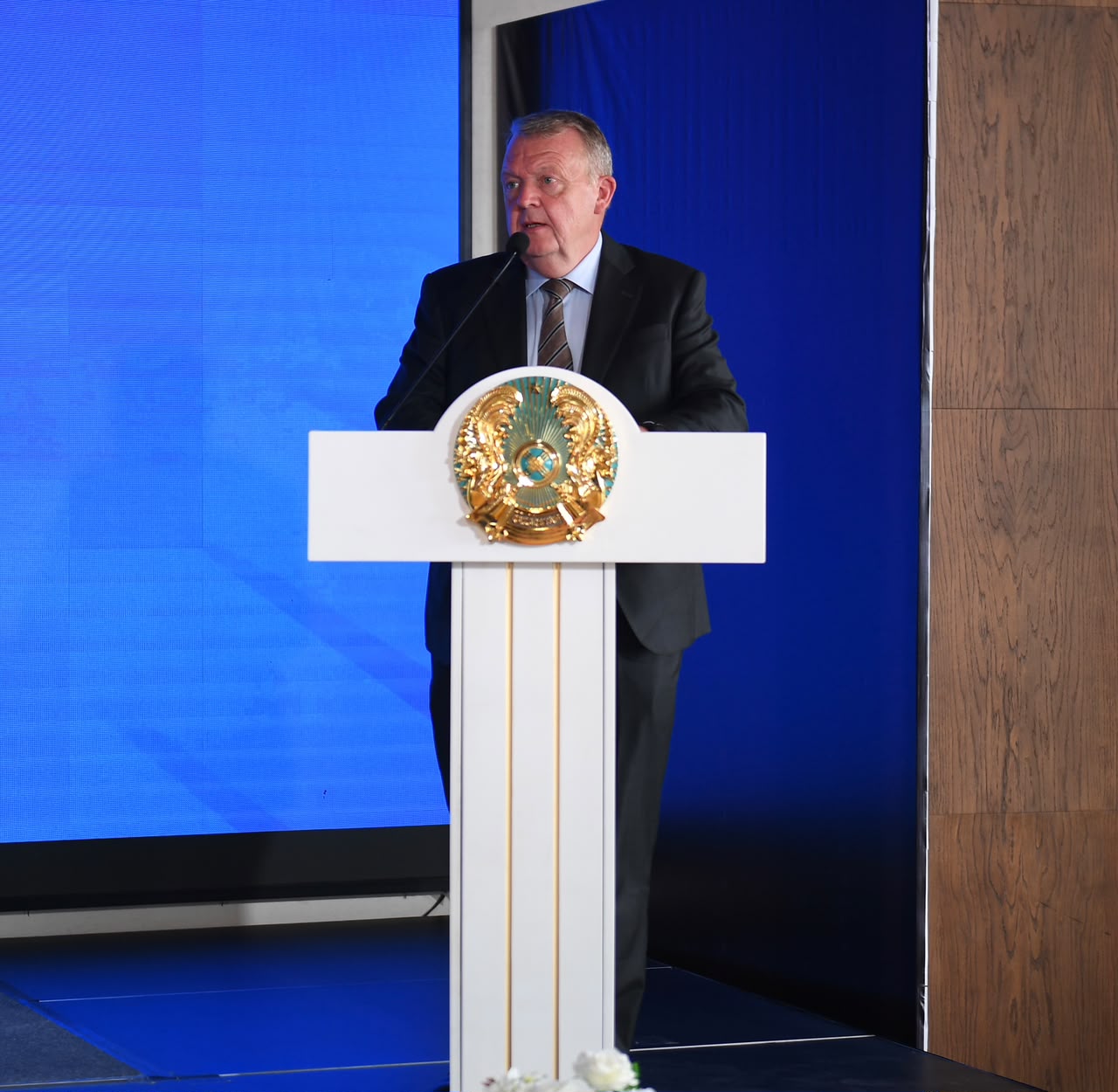 Қазақстан Республикасының гербі бар трибунада сөйлеп тұрған костюм киген ер адам.